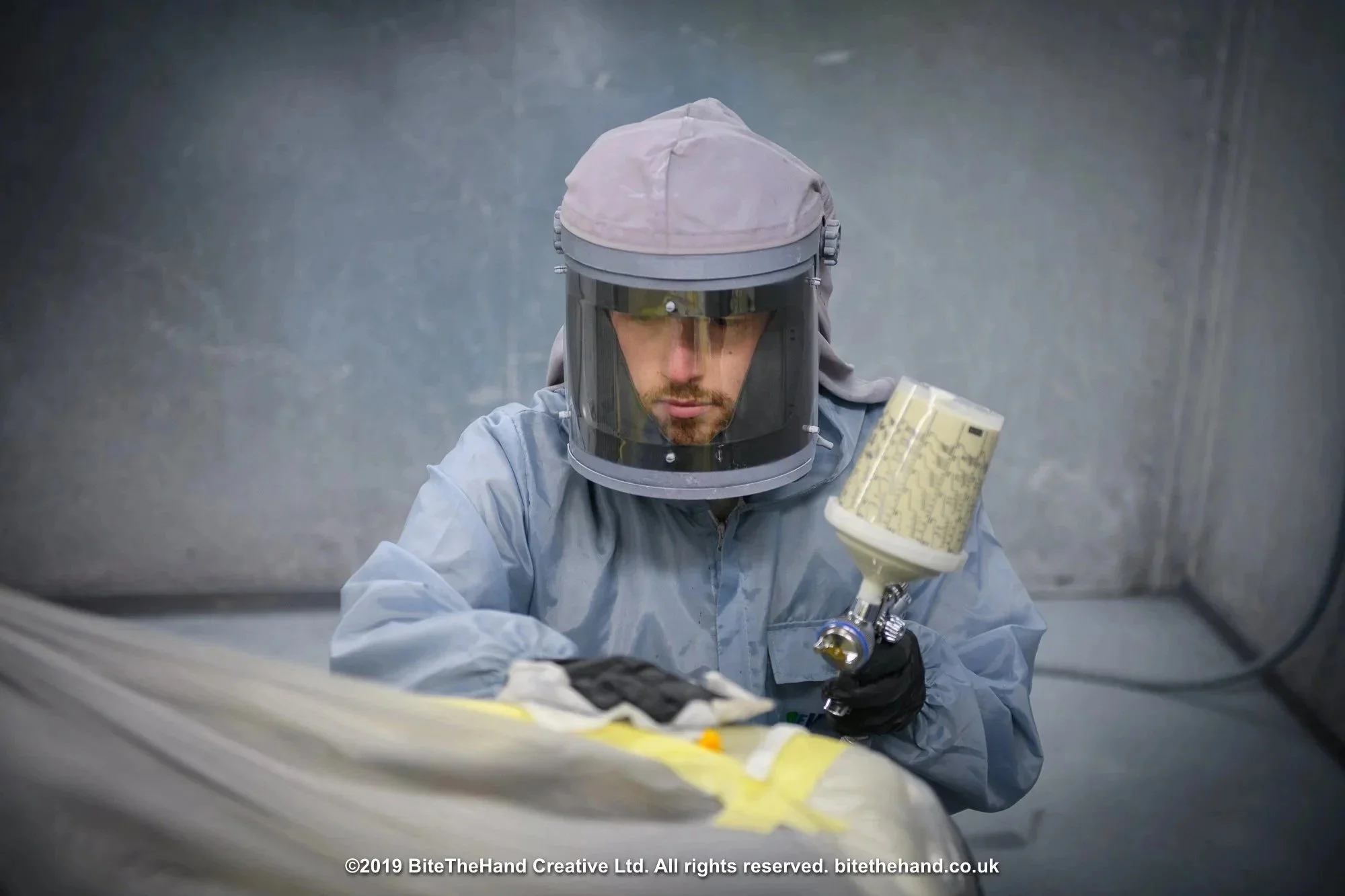 A scientist wearing protective gear, including a hazmat suit and helmet with a face shield, working with a piece of equipment inside a controlled environment.