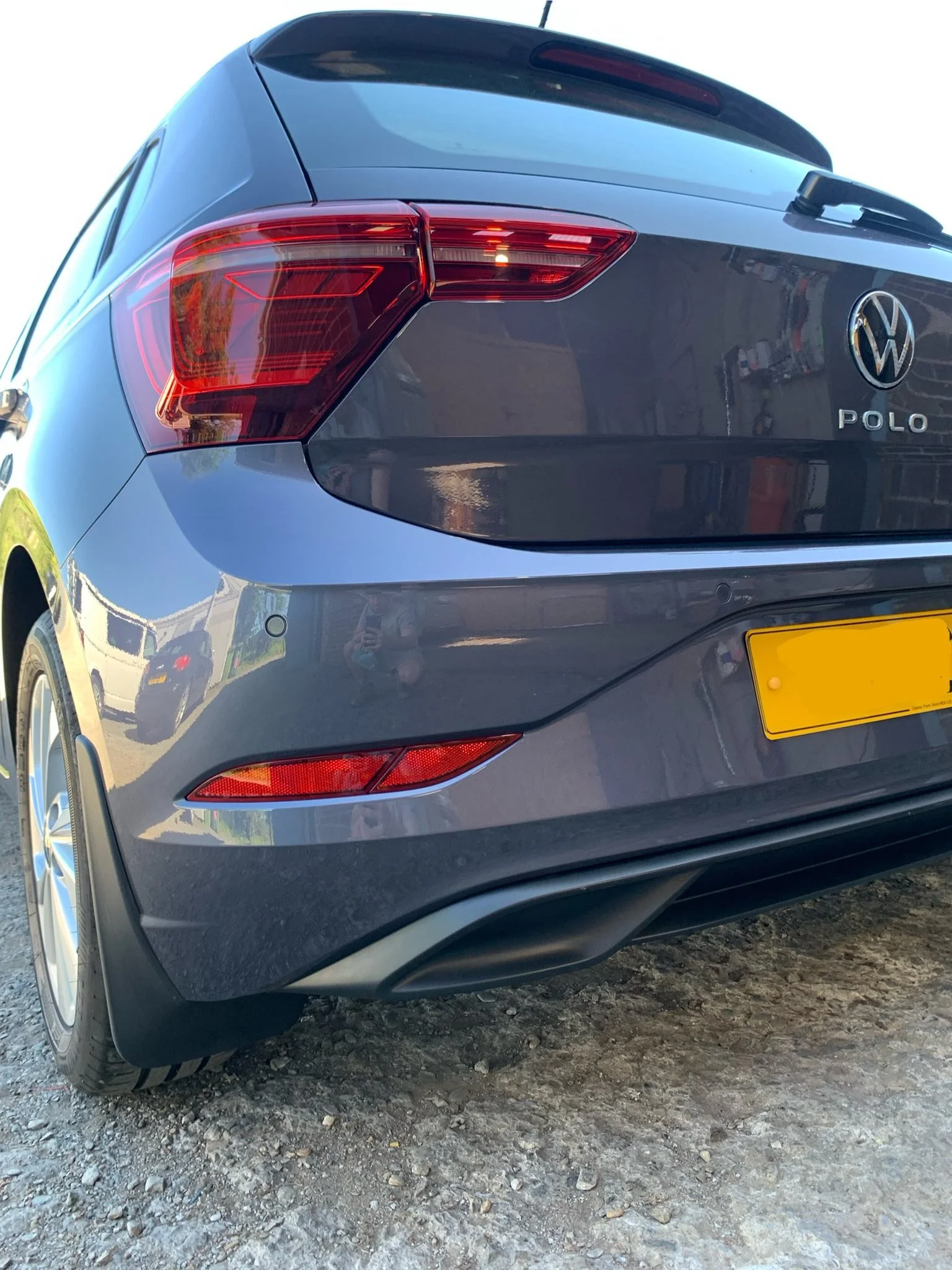 Back of a blue Volkswagen Polo car parked on a gravel surface, showing the taillights, rear bumper, and license plate area.