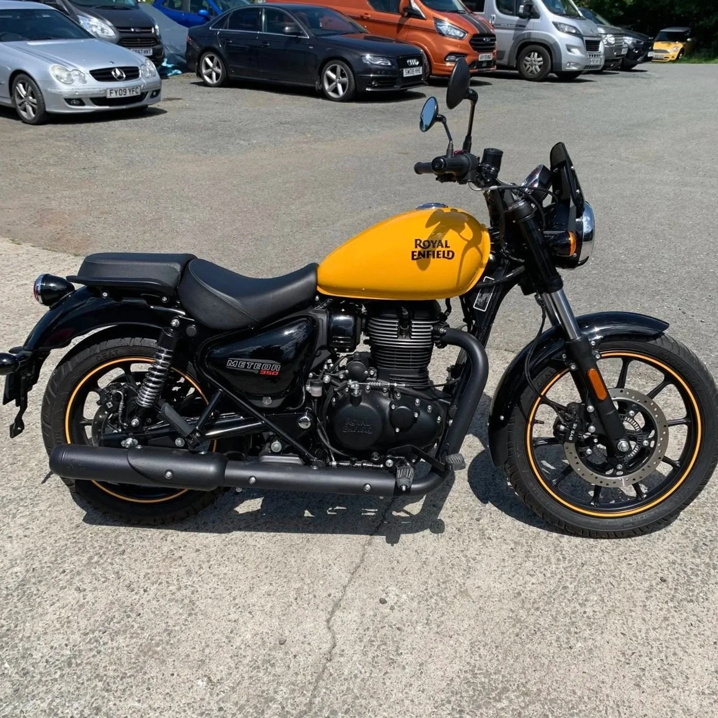 A black and yellow Royal Enfield Meteor 350 motorcycle parked on a gravel lot, with other cars visible in the background.