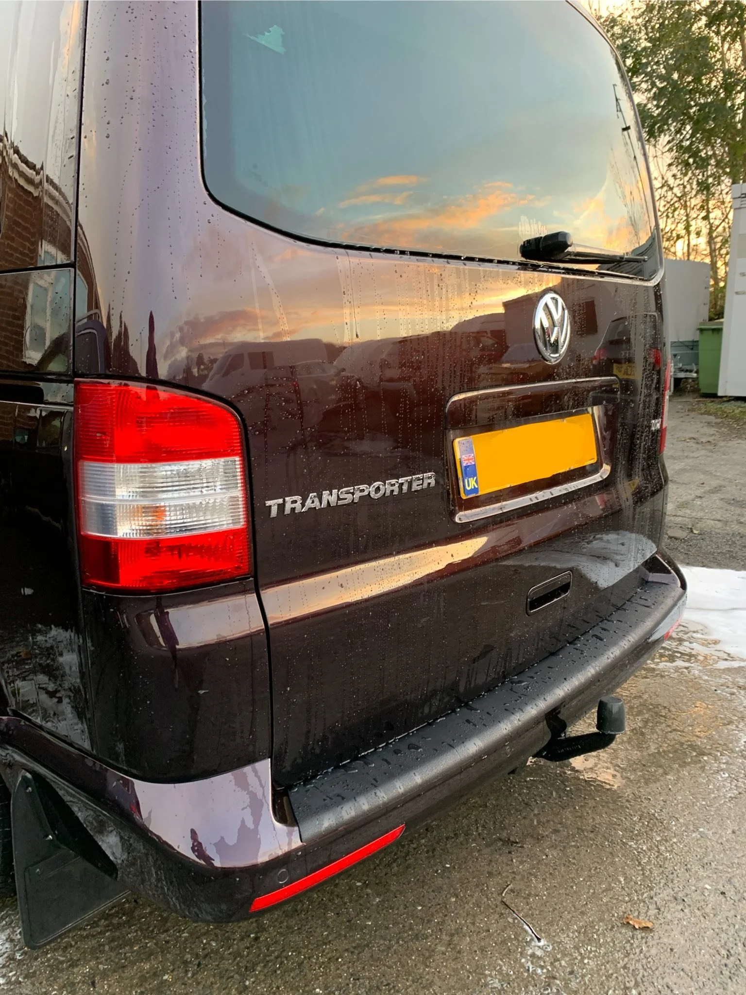 Black Volkswagen Transporter van with rear window reflecting a sunset sky, parked on a wet surface with a green bin and trees in the background.