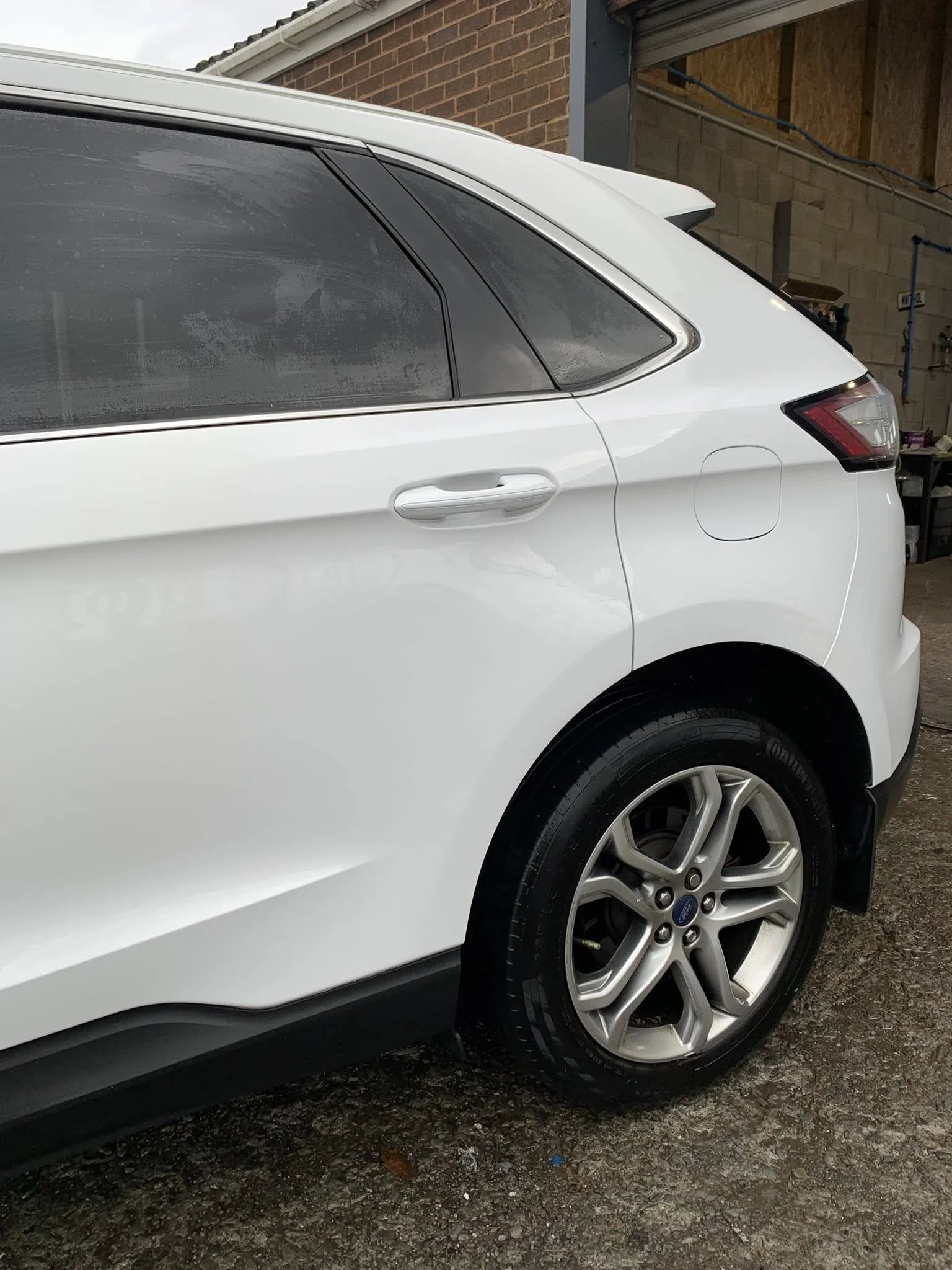 Rear side of a white SUV parked on a gravel driveway, in front of a brick building, with a window and a partially opened garage door.