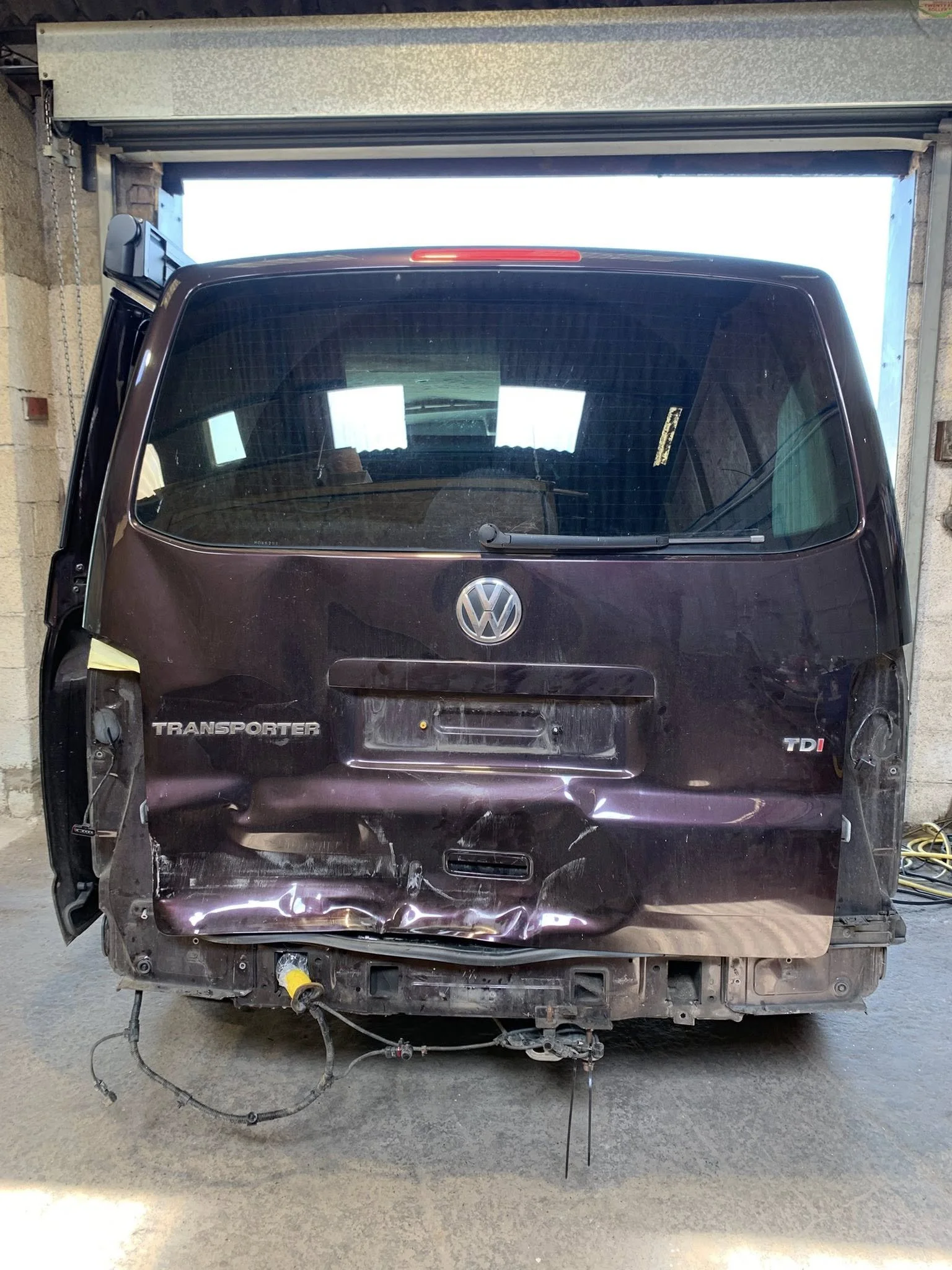 Rear view of a damaged Volkswagen Transporter TDI van with a crushed rear end in an industrial garage.