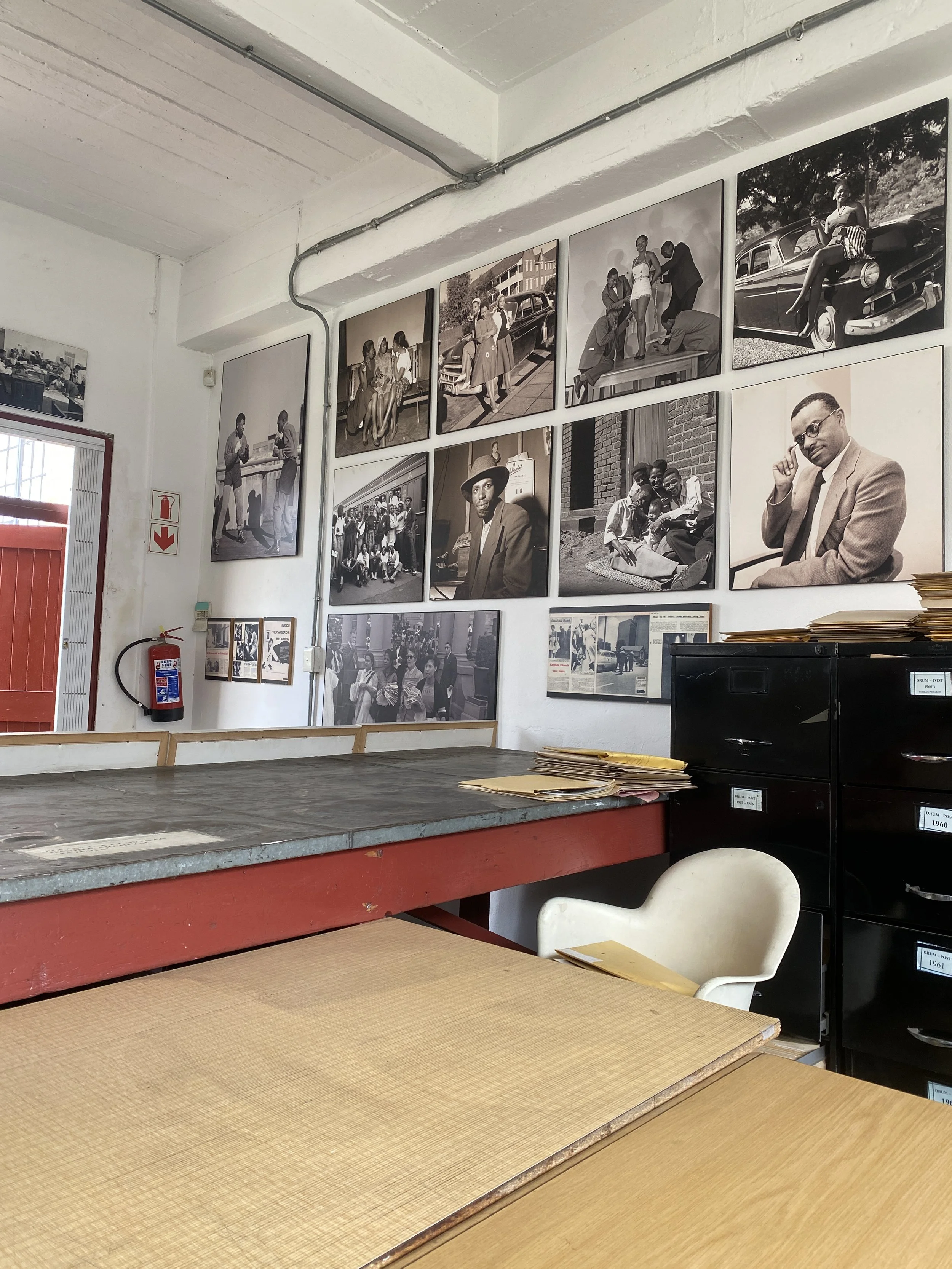 Black and white photographs of people from past decades on a gallery wall in an office space, with a work desk and filing cabinet in the foreground.