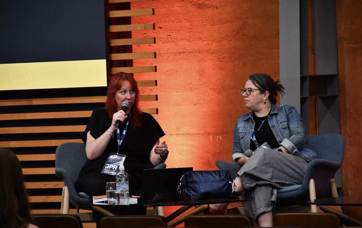 Two women sitting on chairs on a stage with a brick wall background. The woman on the left has red hair and is speaking into a microphone. The woman on the right has dark hair, glasses, and is listening attentively. There are water bottles, a laptop, and a bag on a table in front of them.