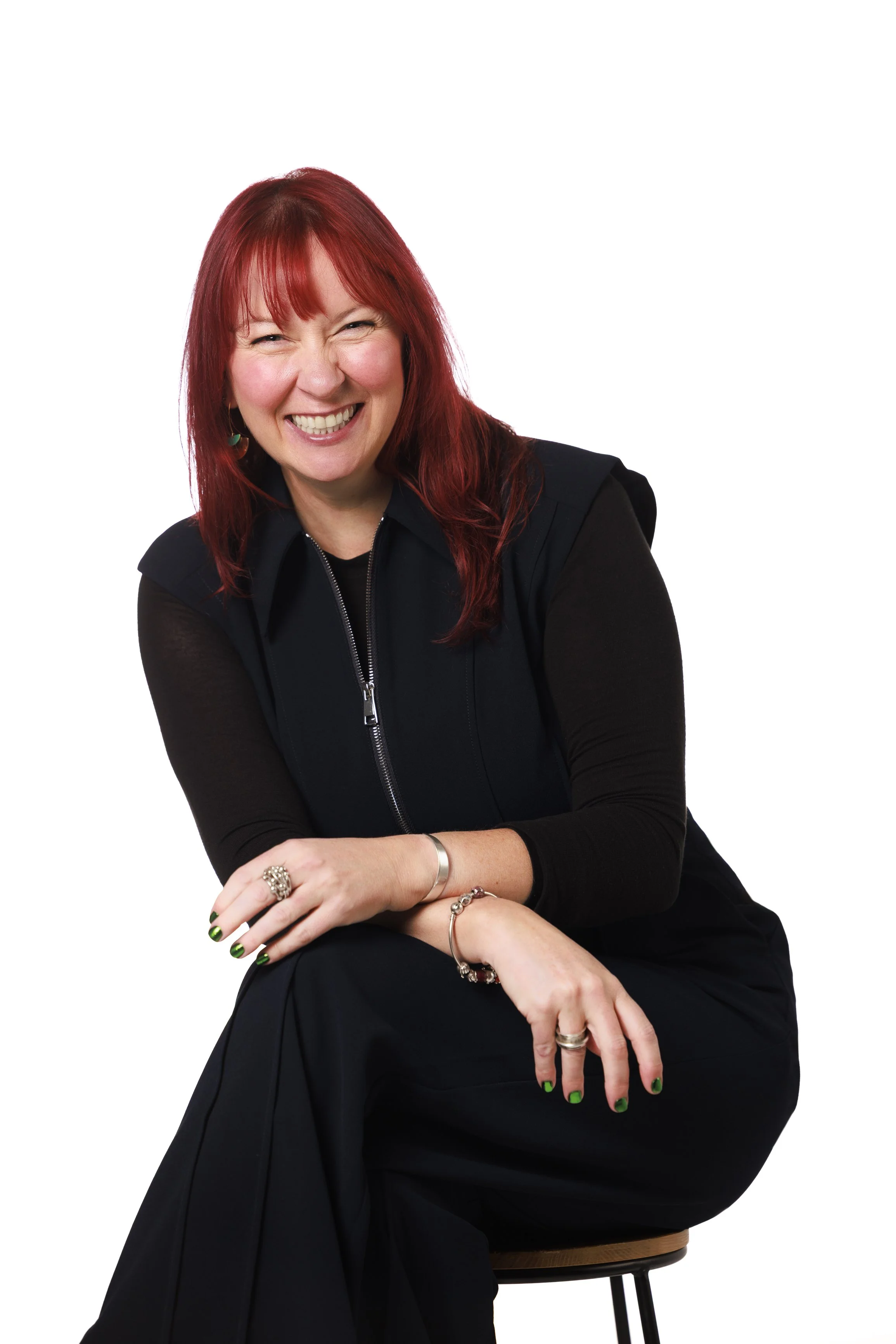 A woman with red hair and a broad smile, sitting on a stool, wearing a black jacket and black dress, with jewelry and painted nails, against a plain white background.