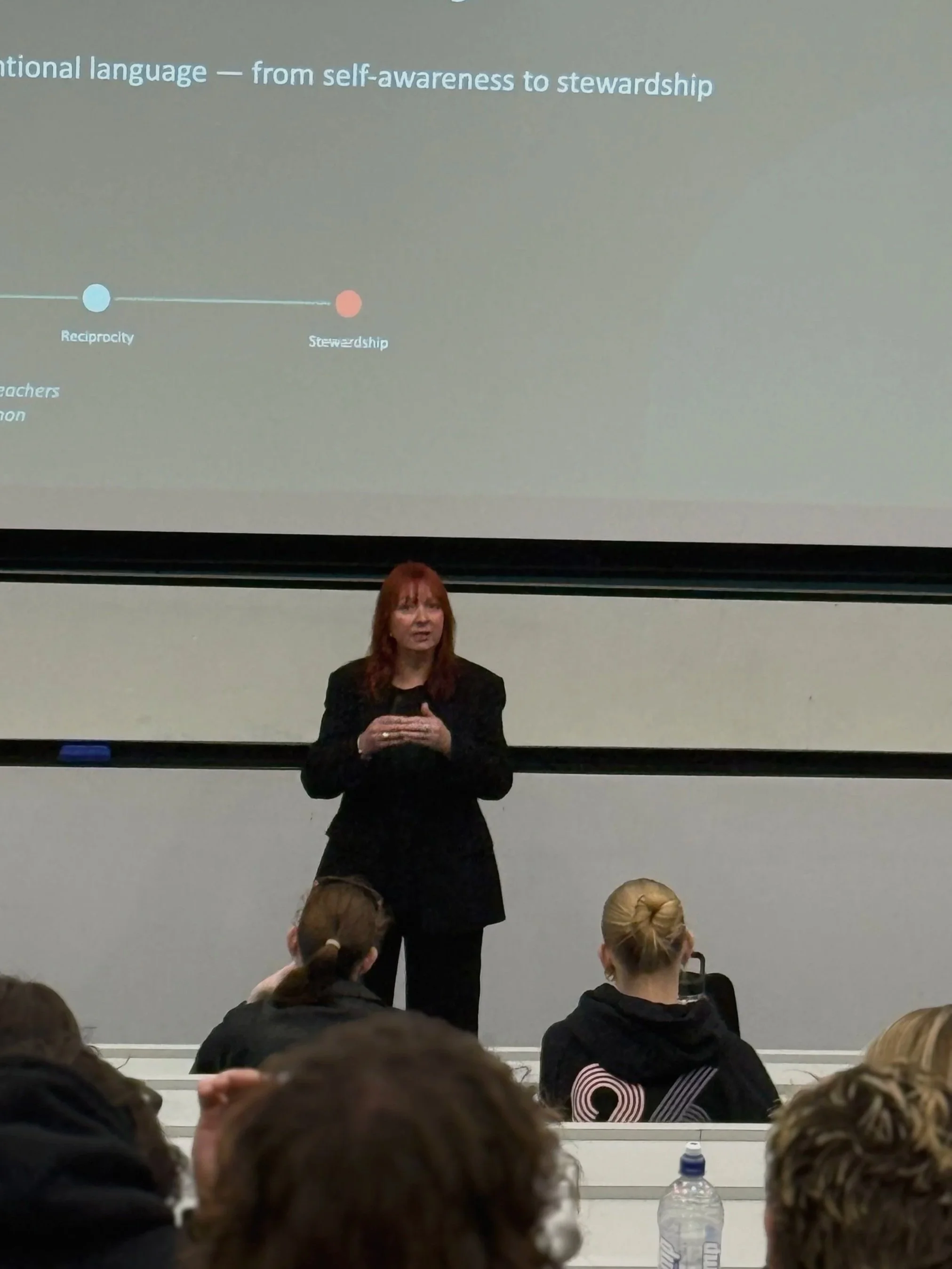 A woman standing in front of a projector screen giving a lecture or presentation to an audience in a classroom or lecture hall.