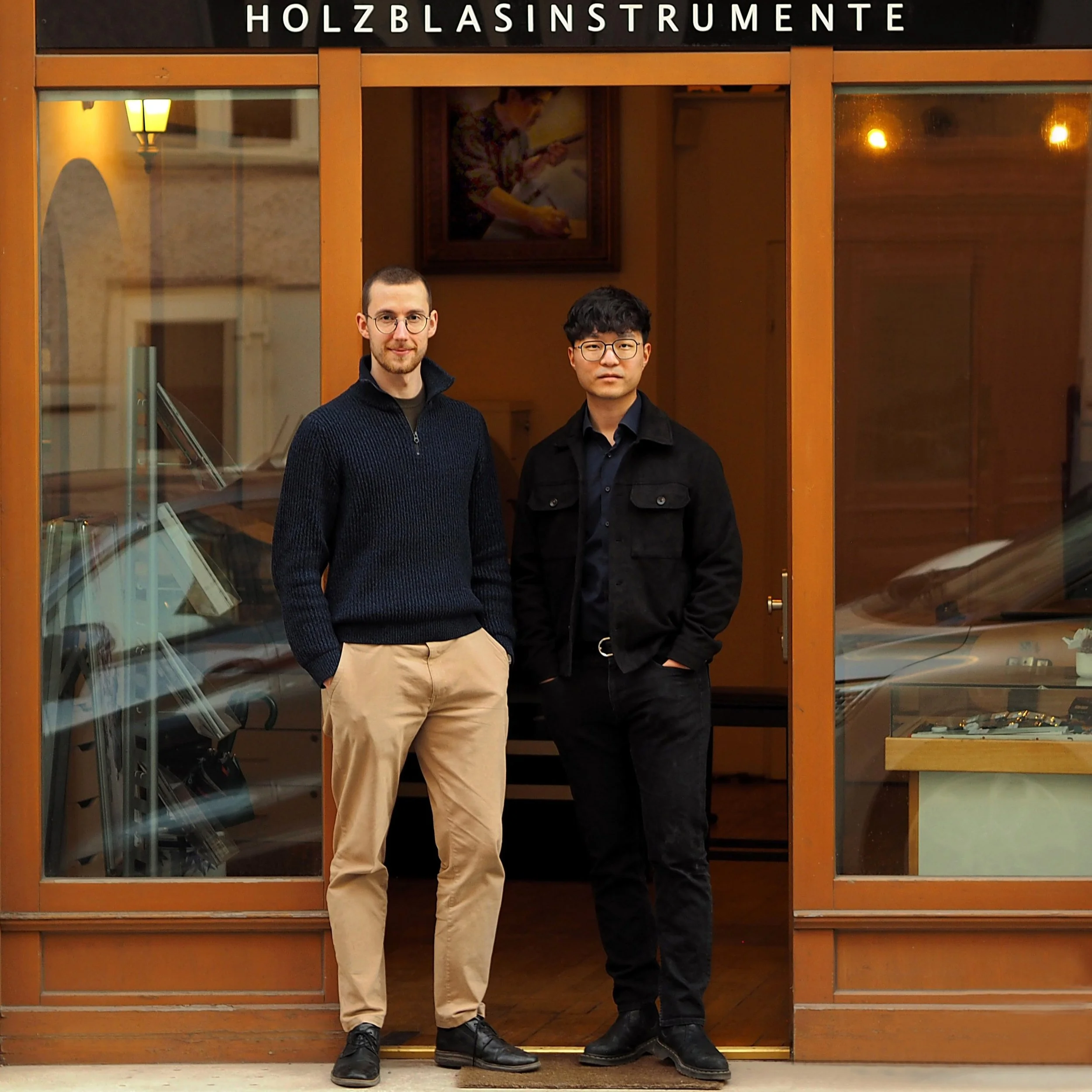 Zwei junge Männer stehen vor einer Glastür mit Holzrahmen, im Hintergrund ein Gemälde, die Tür hat die Aufschrift "Holzblasinstrumente".