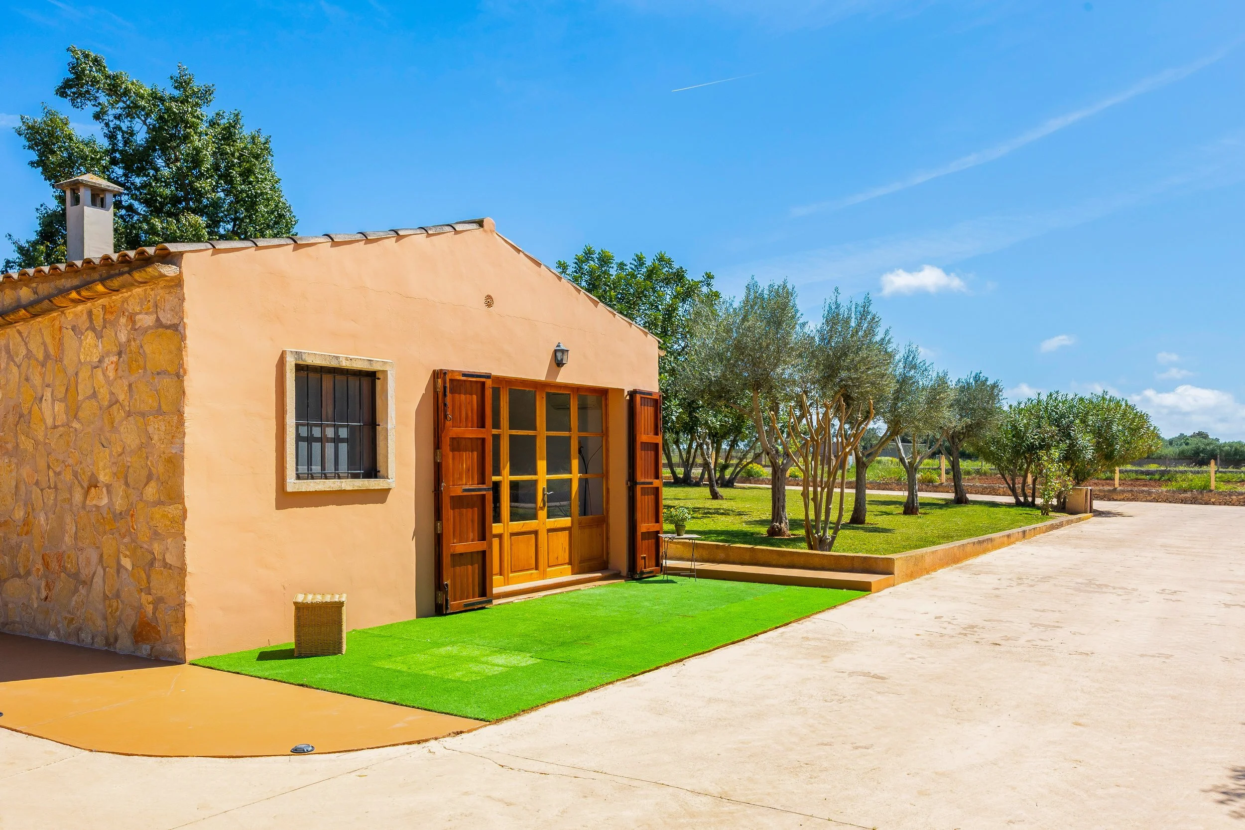 Casa de campo con paredes de color terracota, puertas de madera abiertas, ventana con rejas de hierro y techo de tejas, rodeada de árboles y césped verde, bajo un cielo azul con nubes.