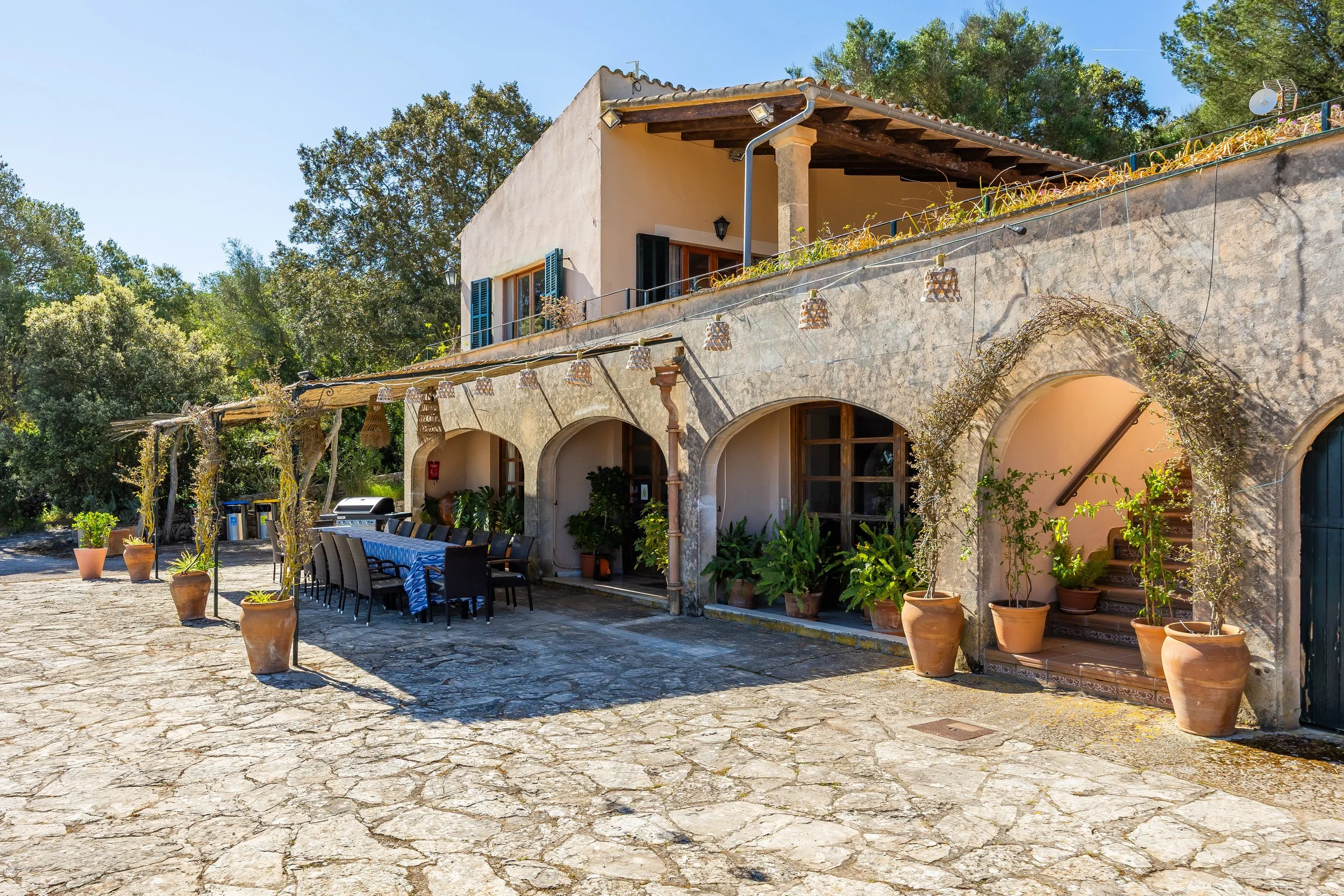Una casa de estilo rústico con paredes de piedra, terraza con ventanas de madera y macetas con plantas, y una mesa larga con sillas en el patio, rodeada de árboles y un cielo azul claro.