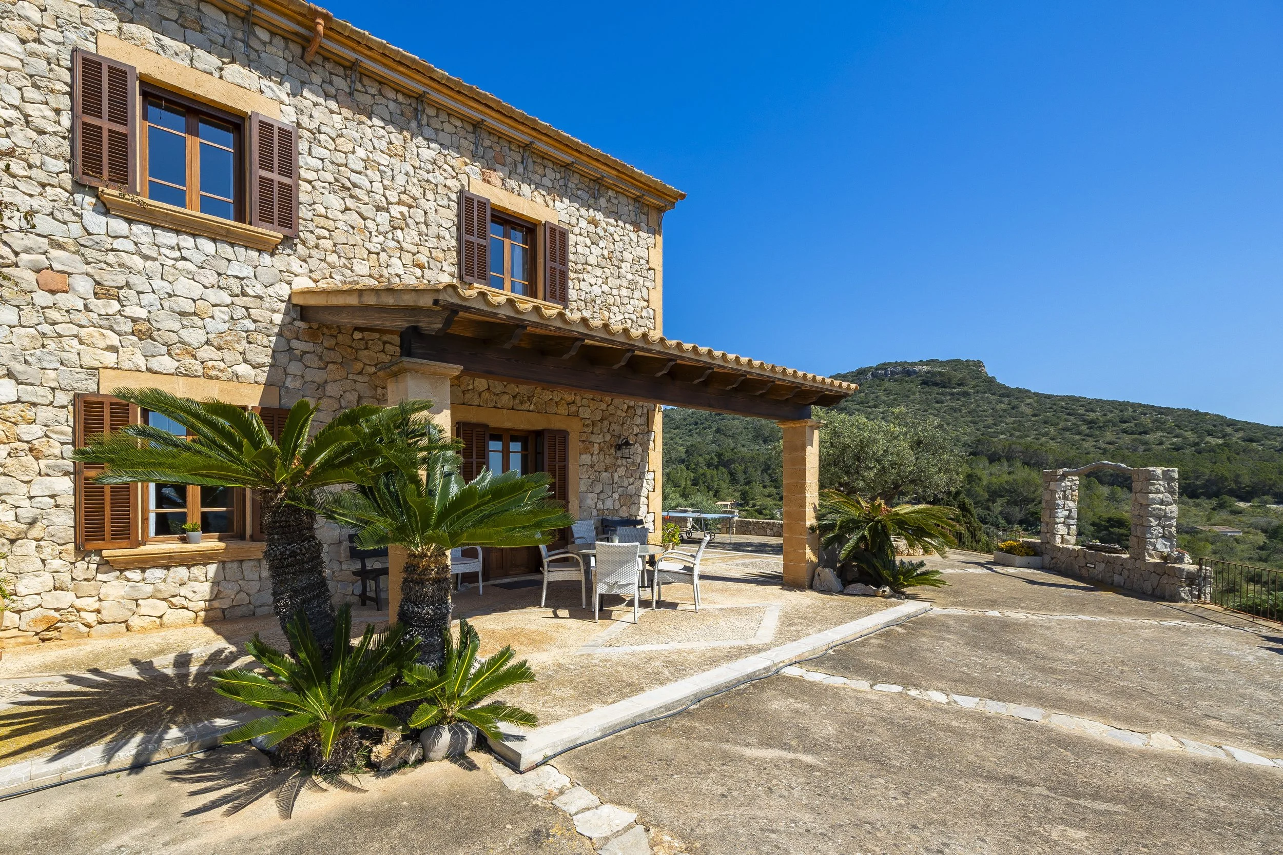 Casa de piedra con ventanas de madera y persianas, terraza con mesa y sillas, palmeras en primer plano, montañas y cielo azul en el fondo.