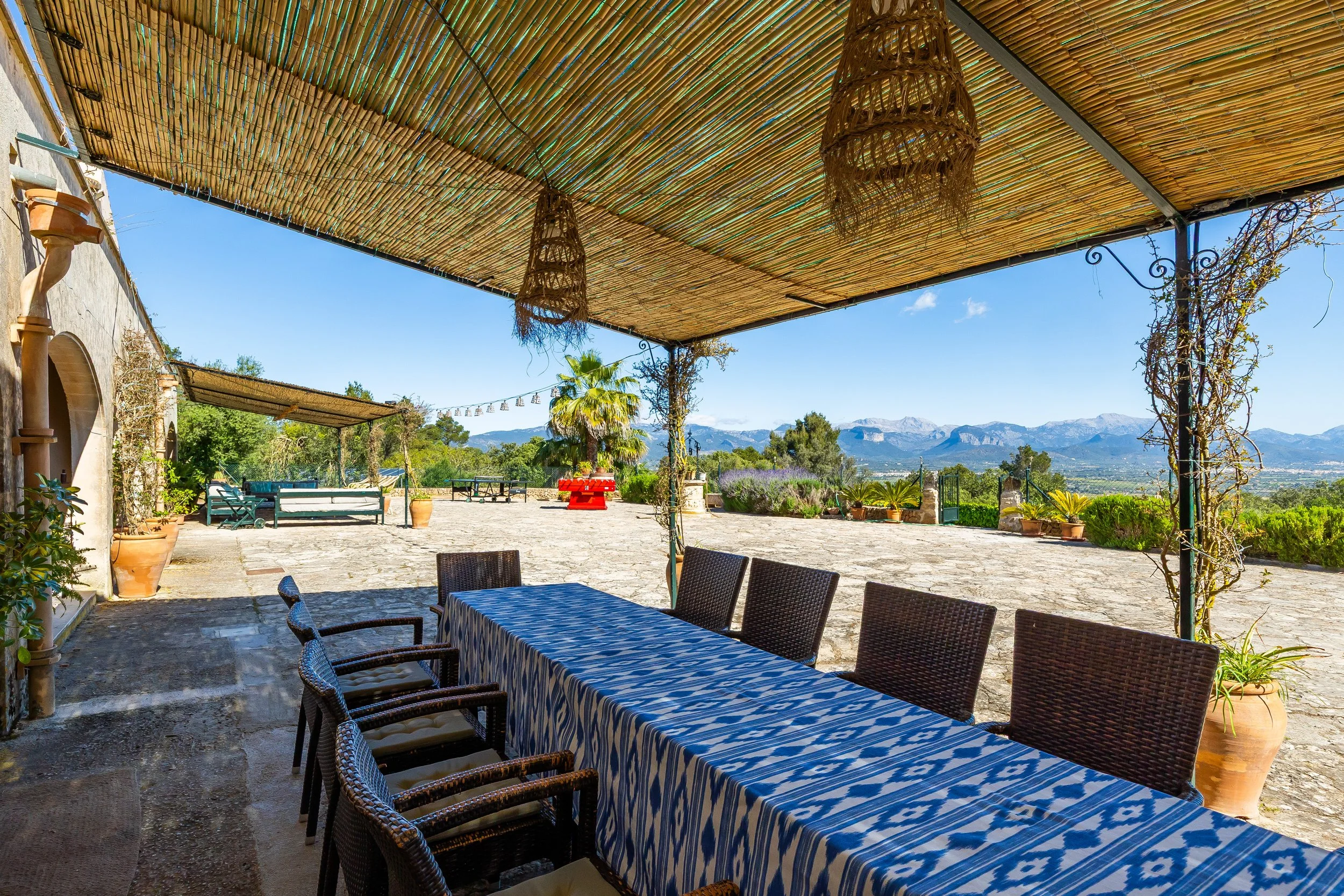 Comedor al aire libre con mesa y sillas bajo cubierta de utensilios de mimbre, con vista a un paisaje montañoso y vegetación diversa en un día soleado.