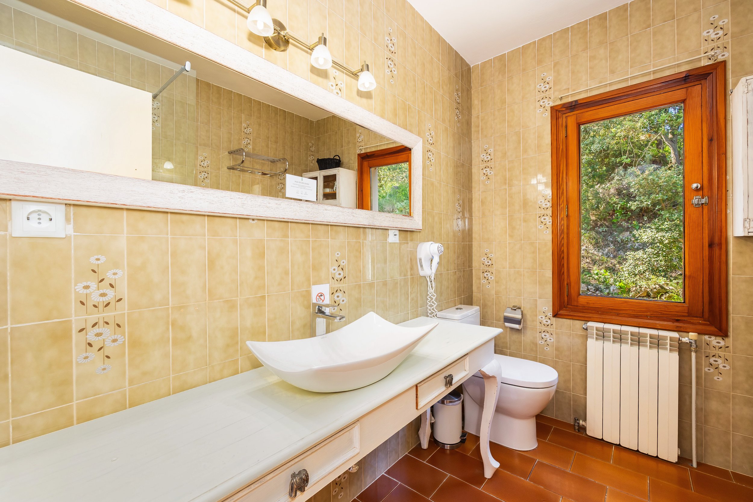 Baño con paredes de azulejos beige con detalles en blanco, ventana con madera que muestra un paisaje de árboles verdes, lavabo de vajilla blanca en una encimera blanca, espejo grande, rubor y accesorios de baño, secador de cabello, inodoro, radiador 