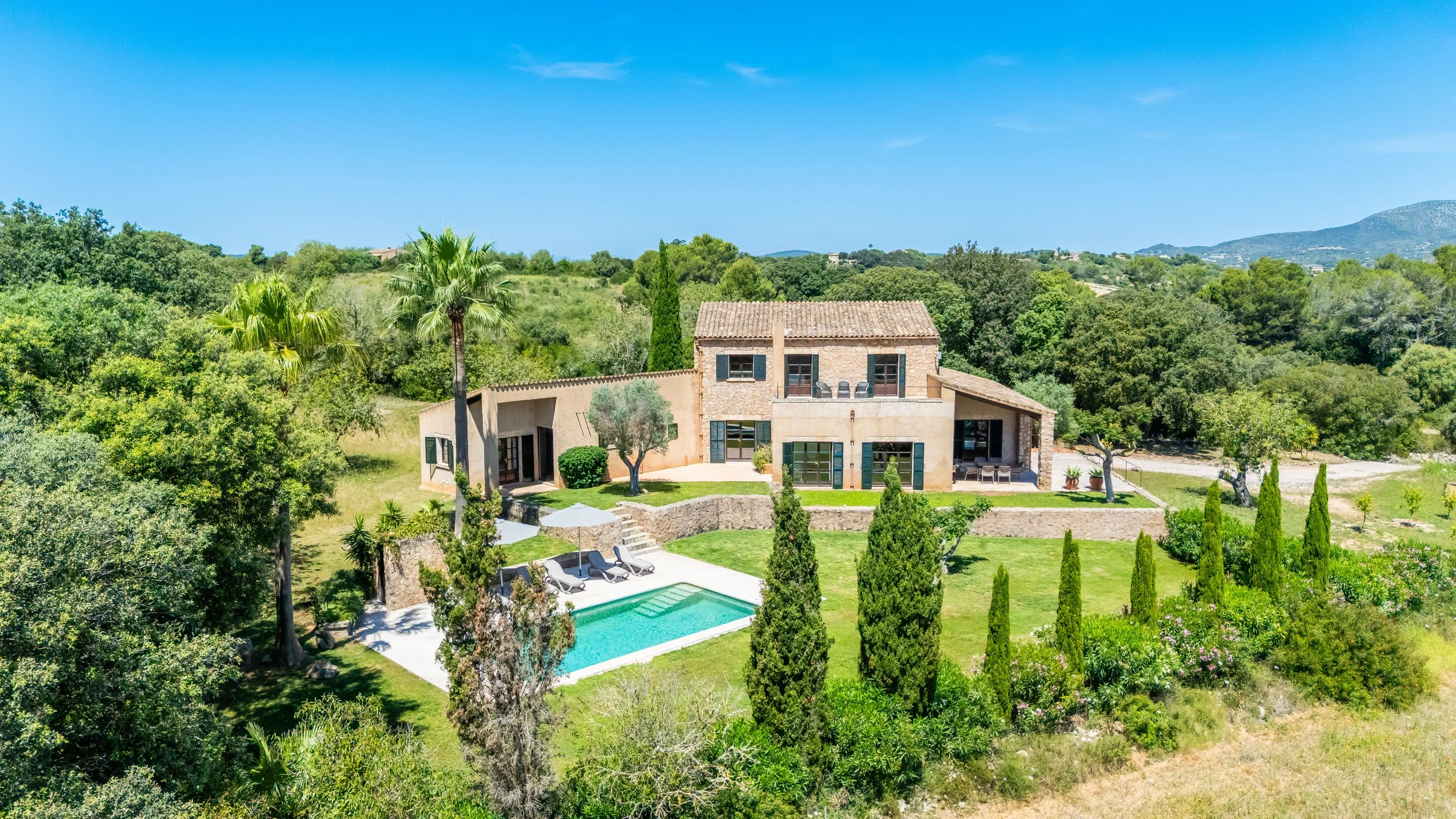 Casa de campo con piscina, rodeada de árboles y vegetación, en un entorno natural con colinas al fondo, bajo cielo despejado.