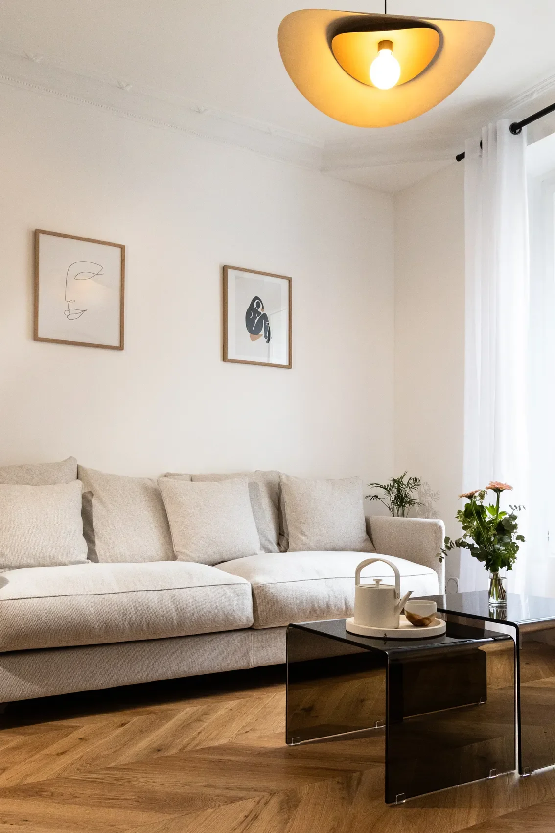 Salon moderne avec canapé beige, deux tableaux minimalistes sur le mur, table basse noire avec théière blanche, vase avec des fleurs et une petite plante, lumière suspendue