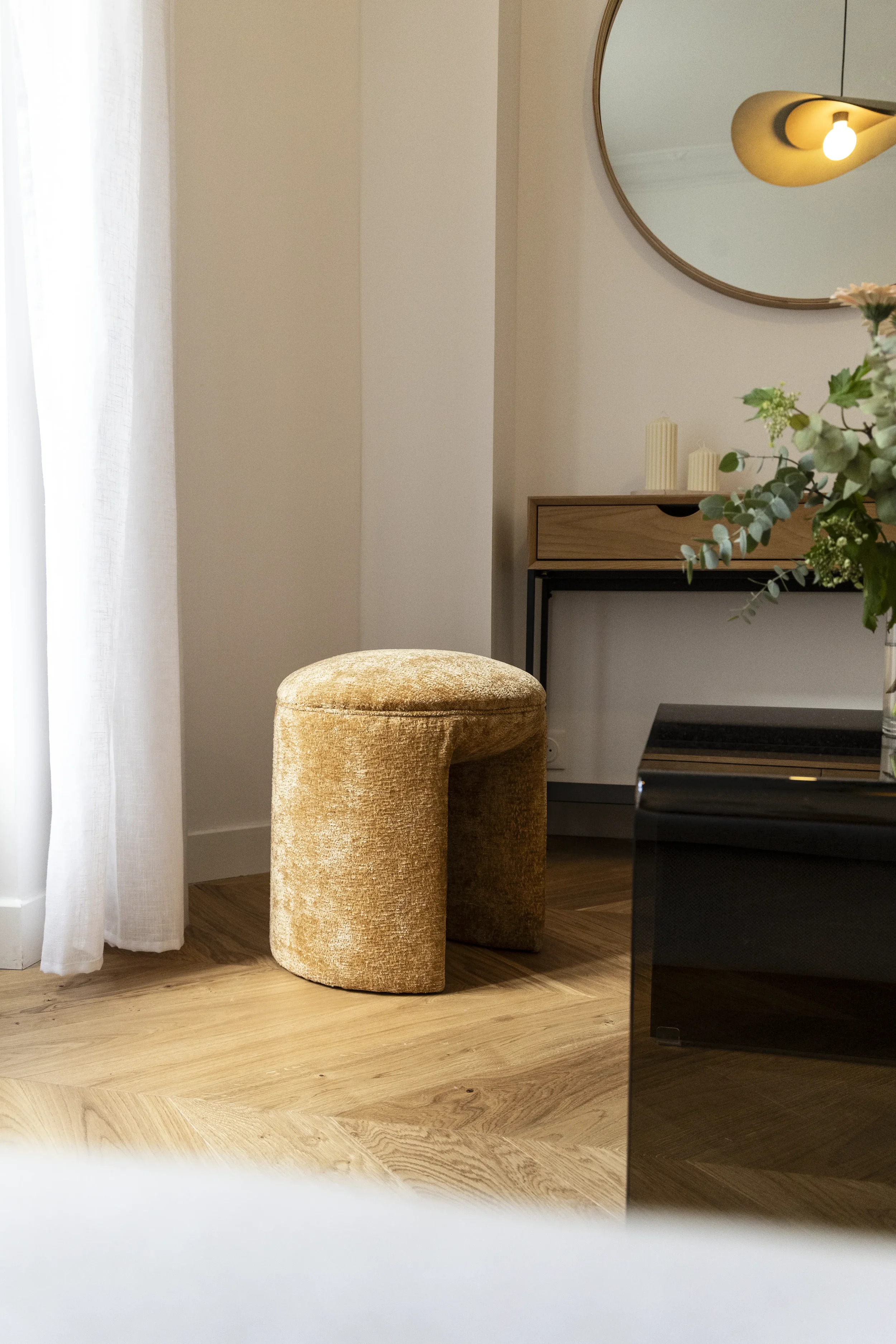 Un intérieur moderne avec un tabouret beige en velours, un meuble en bois avec des bougies, un miroir rond au mur, des plantes vertes et un éclairage chaud, sur un sol en bois.