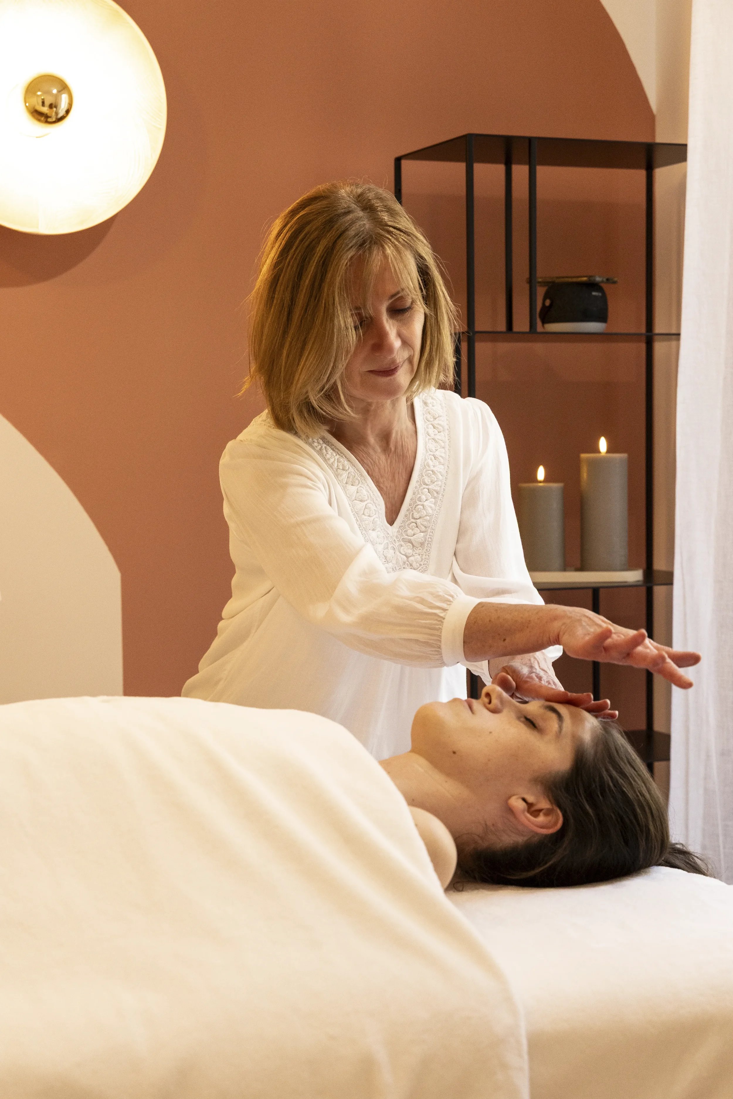 Une femme pratique une séance de massage ou de soins sur une personne allongée, dans une ambiance calme et chaleureuse avec des bougies et une décoration moderne.