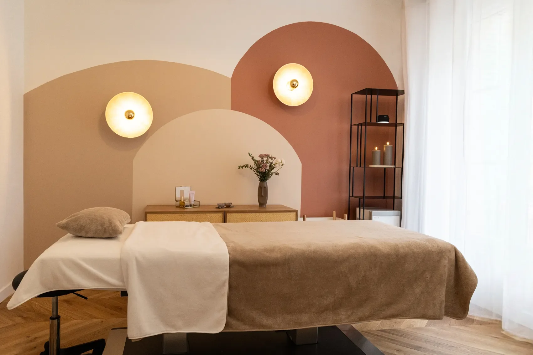 Une pièce de spa ou de massage avec un lit ergonomique recouvert d'une couverture beige, une taie d'oreiller grise, un fond de mur avec des couleurs pastel et des appliques murales lumineuses, une étagère noire avec des bougies et un bouquet de fleurs dans un vase, une grande fenêtre avec des rideaux blancs.