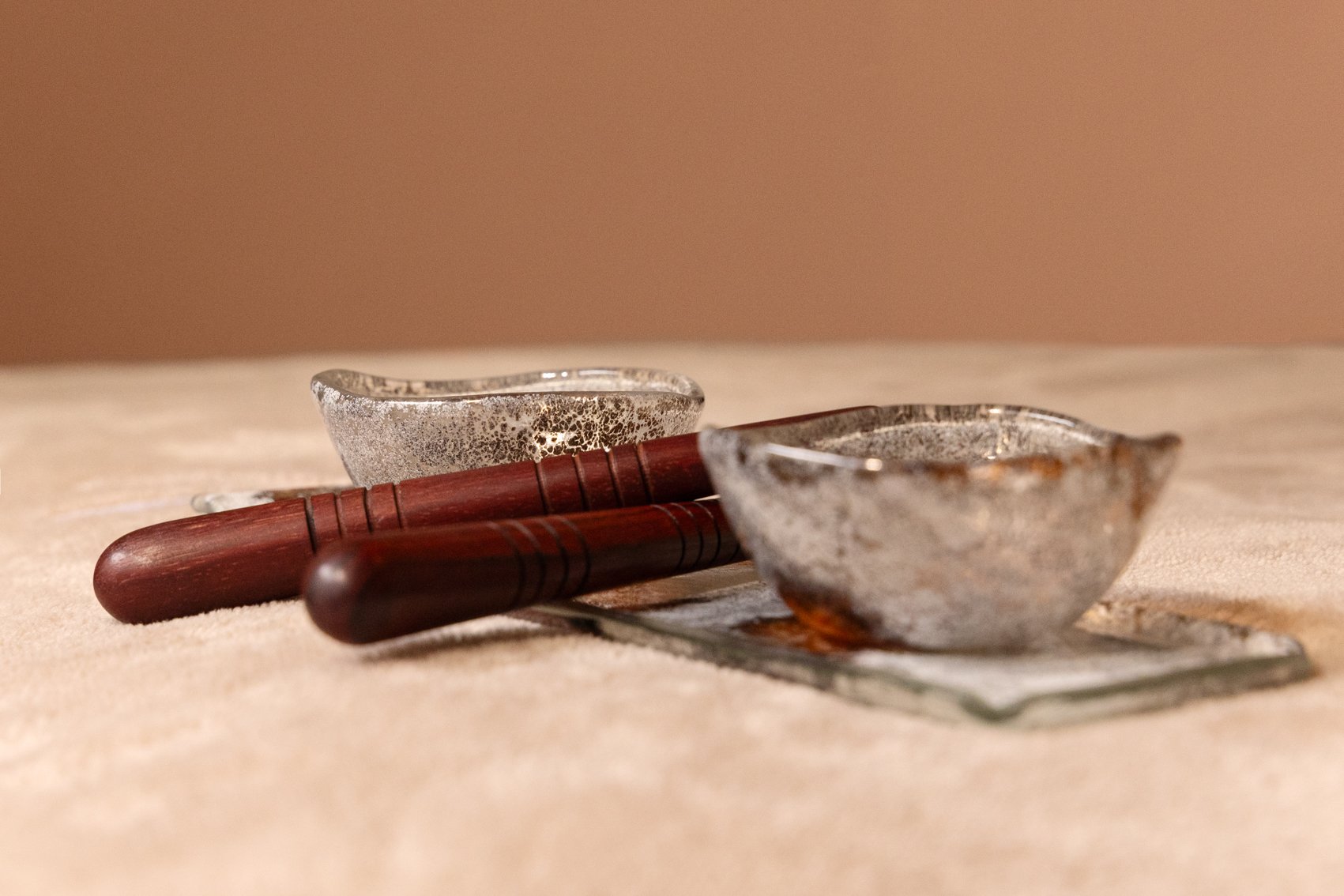 Deux bols en verre avec une texture métallique, deux bâtons en bois rouges, et un tapis en tissu beige sur une surface en bois.