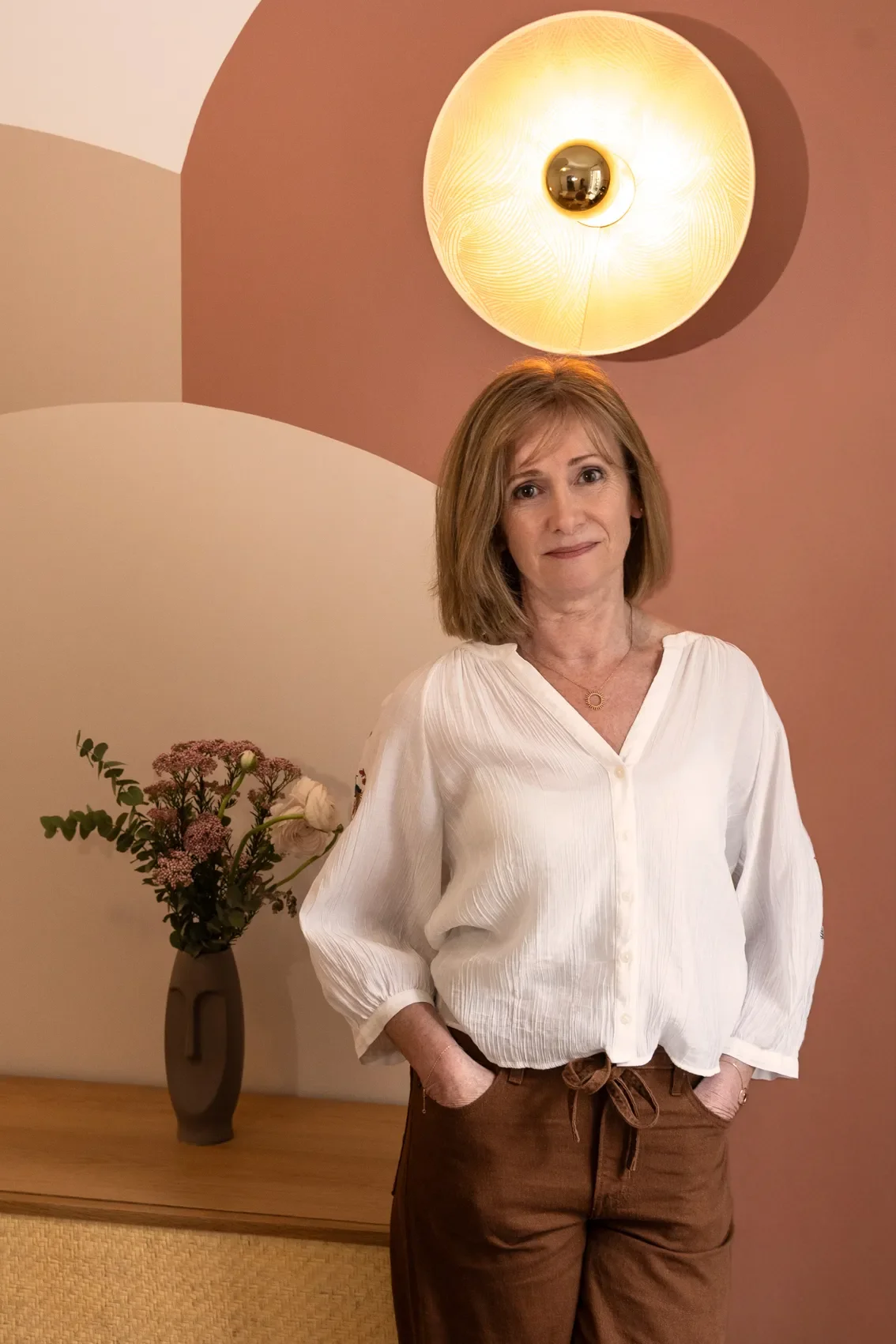 Une femme souriante avec des cheveux bruns courts, portant une chemise blanche et un pantalon marron, debout dans une pièce intérieure. En arrière-plan, il y a une table avec un vase contenant des fleurs et un mur avec un luminaire rond.