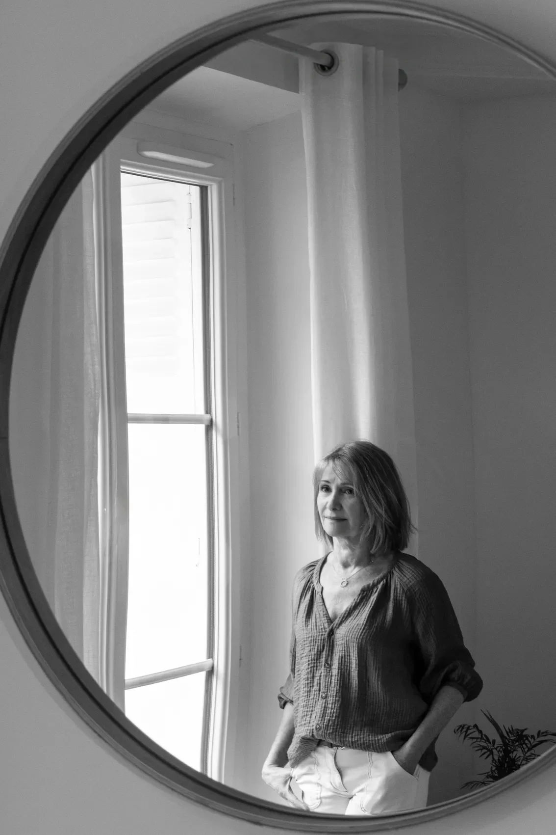 Photo en noir et blanc d'une femme avec des cheveux mi-longs, portant une chemise à manches longues, vue dans un miroir rond, avec une fenêtre derrière elle.