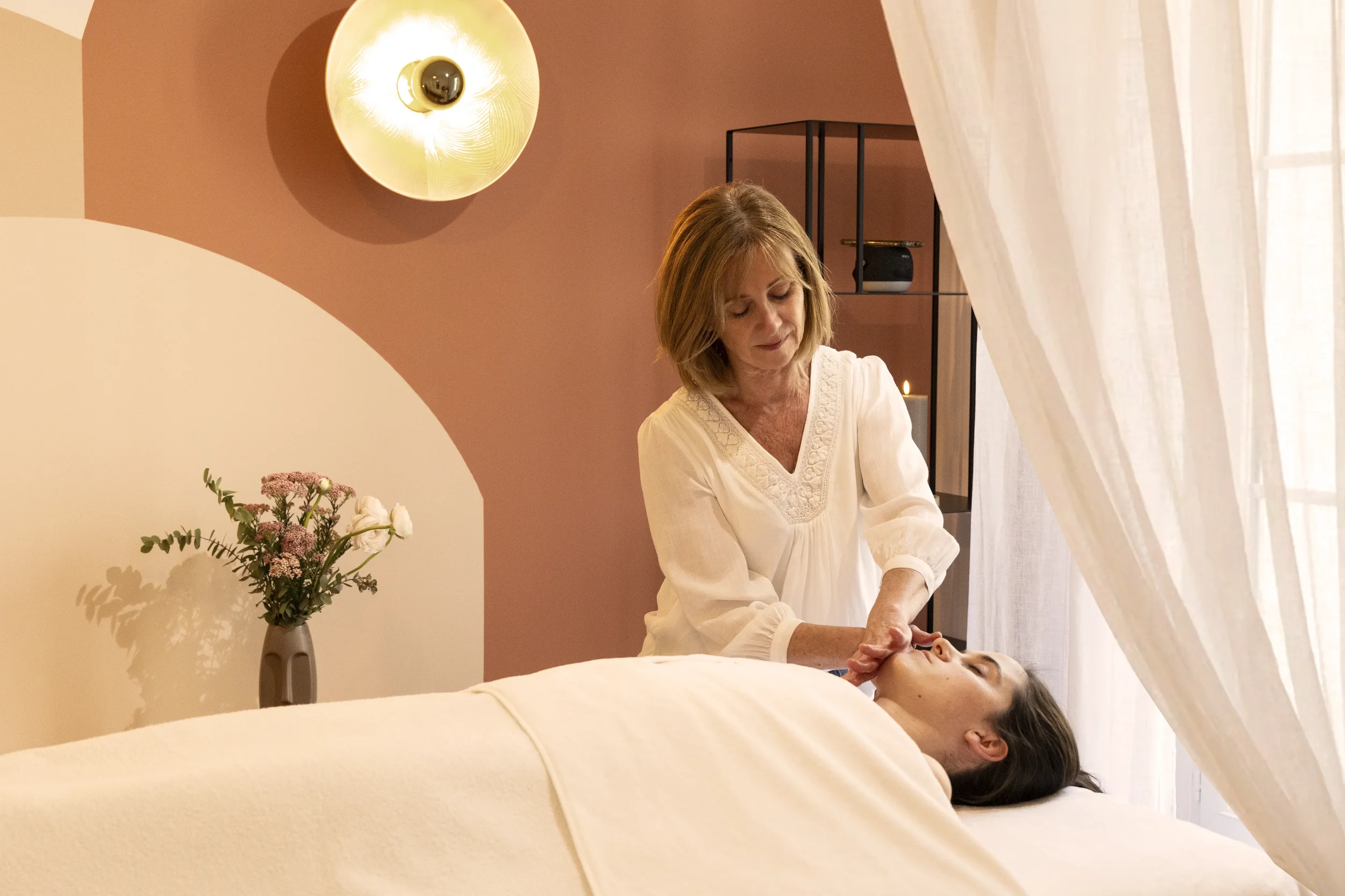 Une femme fait un massage à un homme allongé sur une table de massage dans une pièce cosy avec une décoration moderne, des fleurs dans un vase et un éclairage chaud.