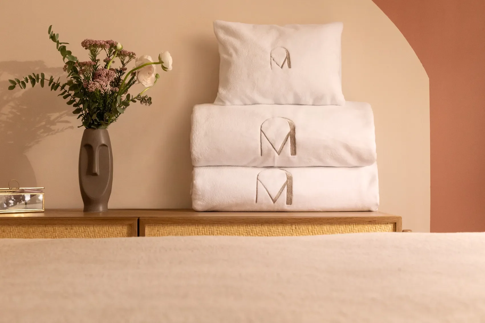 Une pièce décorative avec une vase contenant des fleurs et trois serviettes blanches pliées sur une commode en bois, avec un mur beige et une arche au fond.