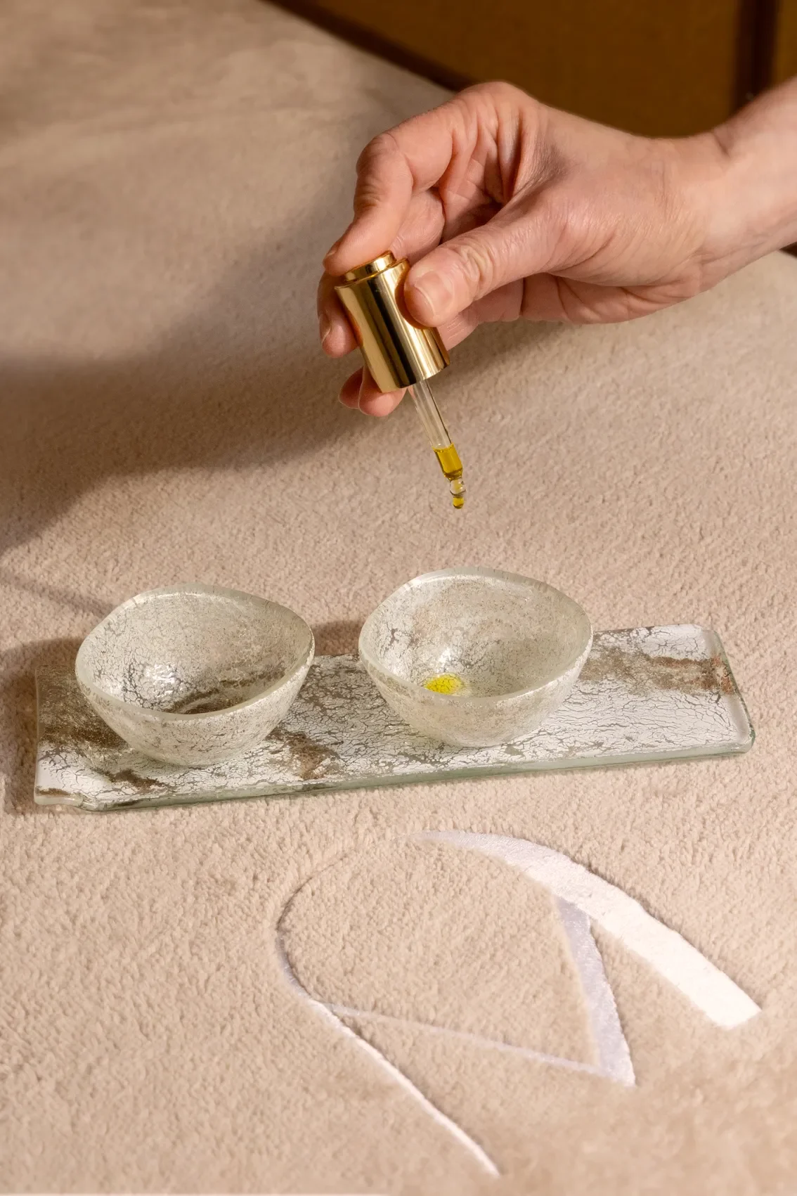 Une main verse un liquide jaune dans un petit bol en verre sur une table en céramique.