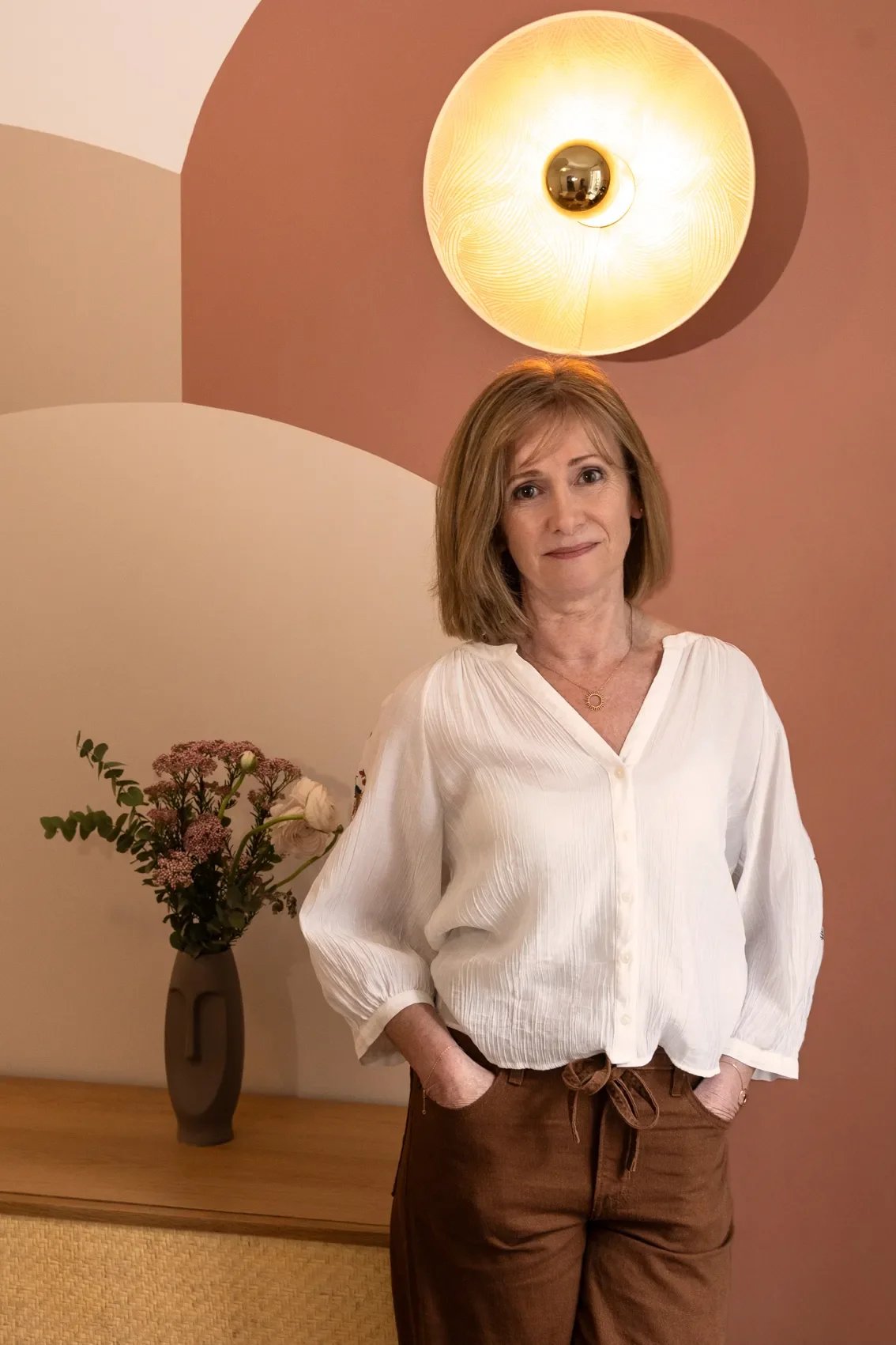 Une femme aux cheveux bruns, porte un chemisier blanc et un pantalon marron, se tient dans une pièce avec un mur coloré en arrière-plan, un luminaire rond jaune brillant suspendu au plafond, et une table avec un vase de fleurs derrière elle.