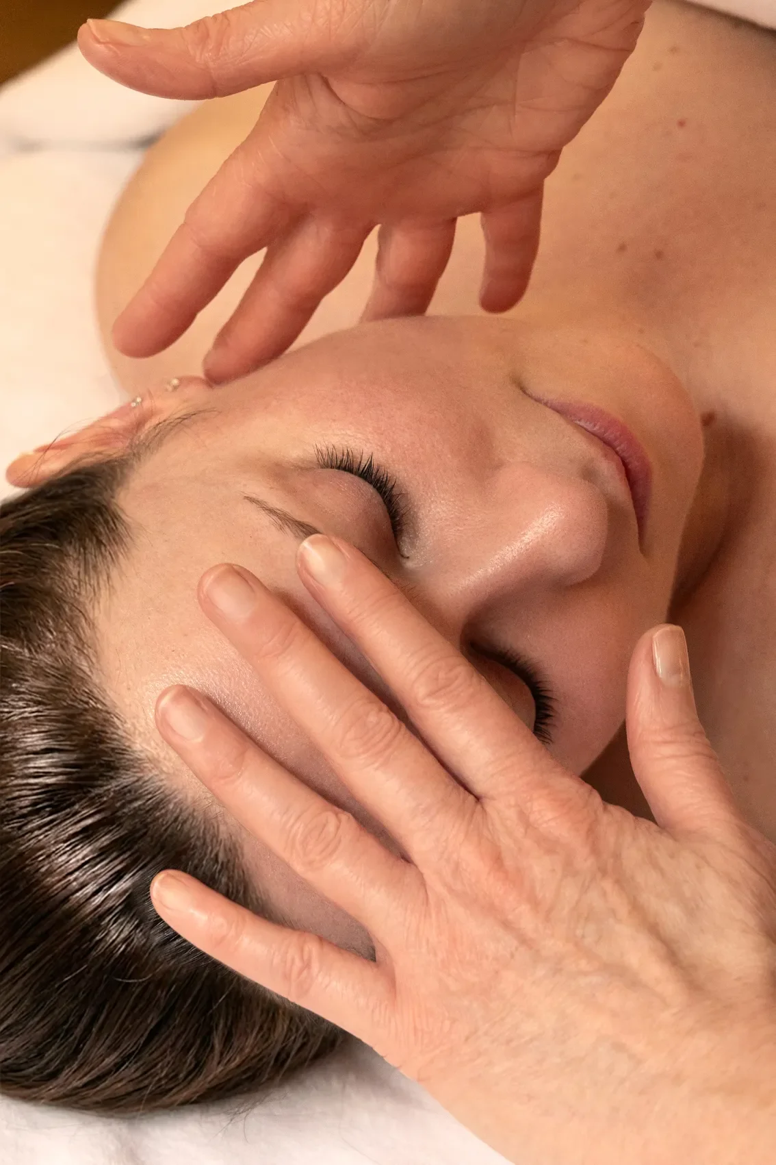 Une personne reçoit un massage facial, allongée avec les yeux fermés, tandis que deux mains massent doucement son visage.