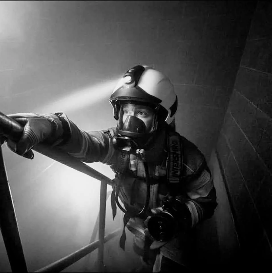 Firefighter in helmet and protective gear using a hose, water spraying from the nozzle, inside a smoke-filled room.