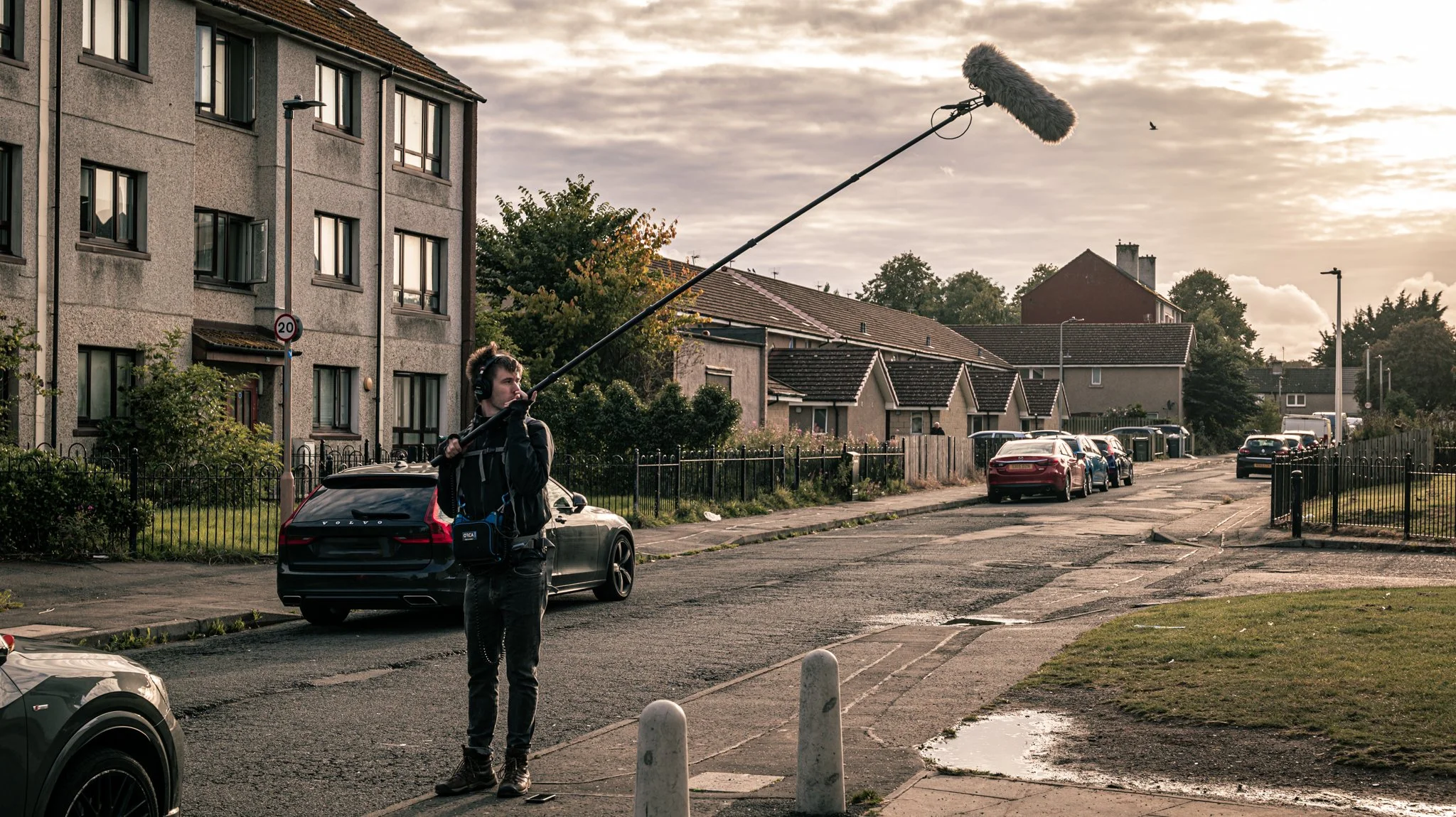 A person holding a boom microphone stands on a residential street, capturing audio as cars are parked along the sidewalk, with houses and trees in the background during sunset.