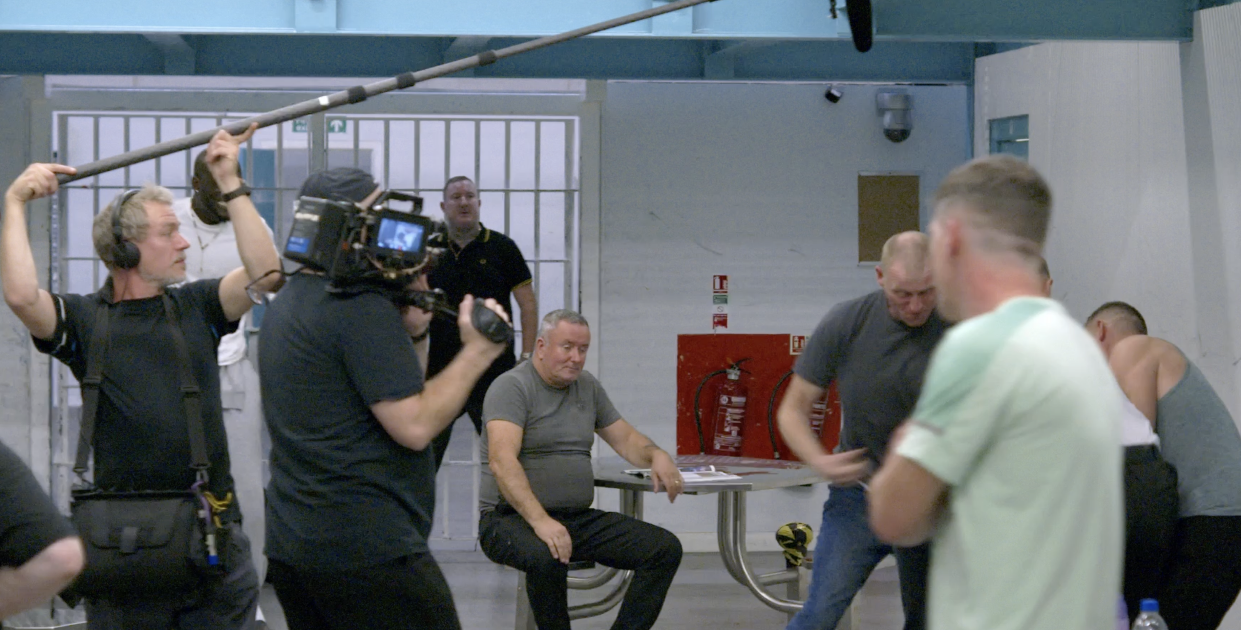 Filming crew working in an indoor gym, with people participating in a physical activity, some seated and some moving, under bright lighting with a fire extinguisher and emergency signs on the wall.