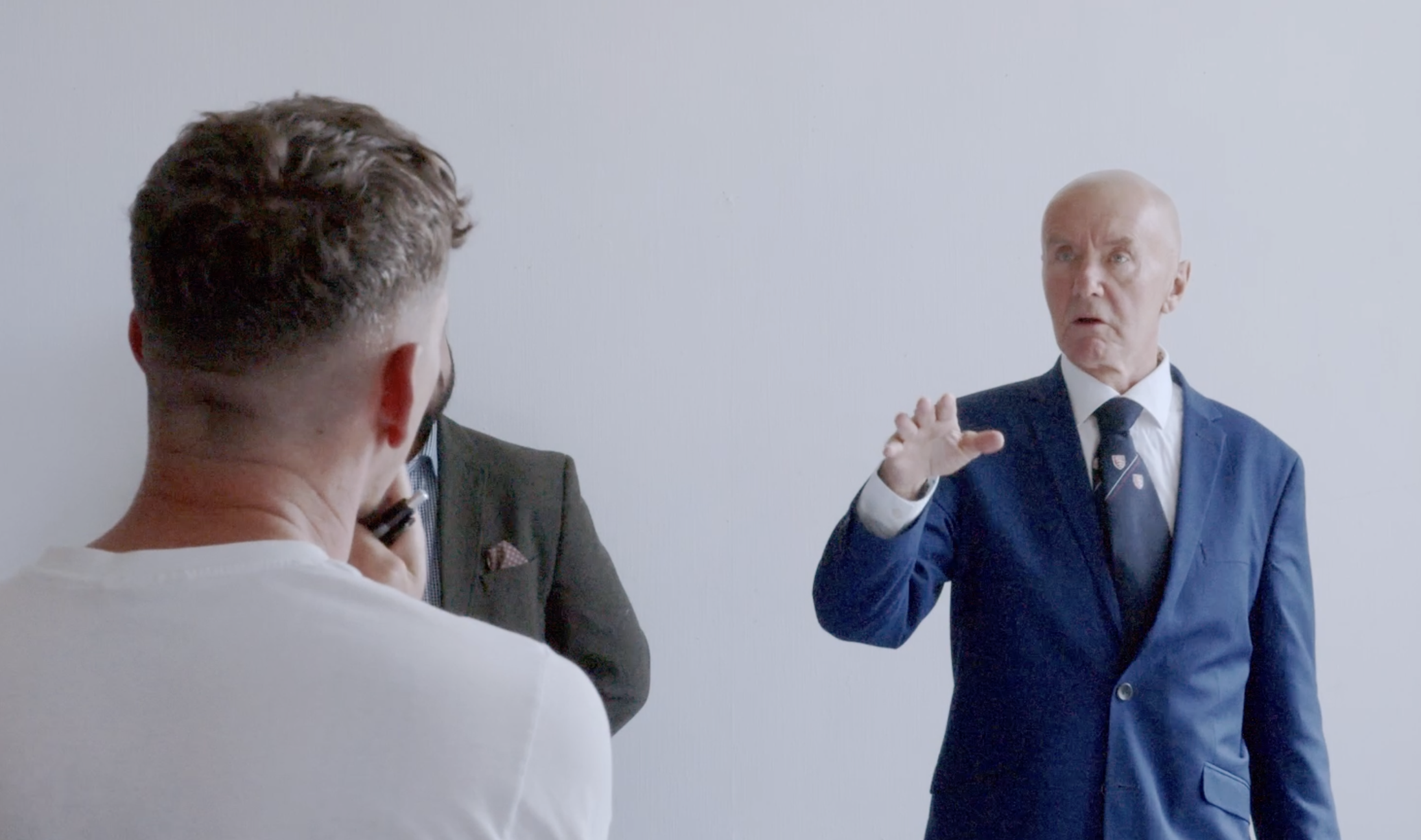 Scene of three men engaged in conversation against a plain light gray background. One man is facing away from the camera with brown hair and a white shirt, while the other two men are facing towards the camera, dressed in formal suits, with the older man making a gesture with his hand.