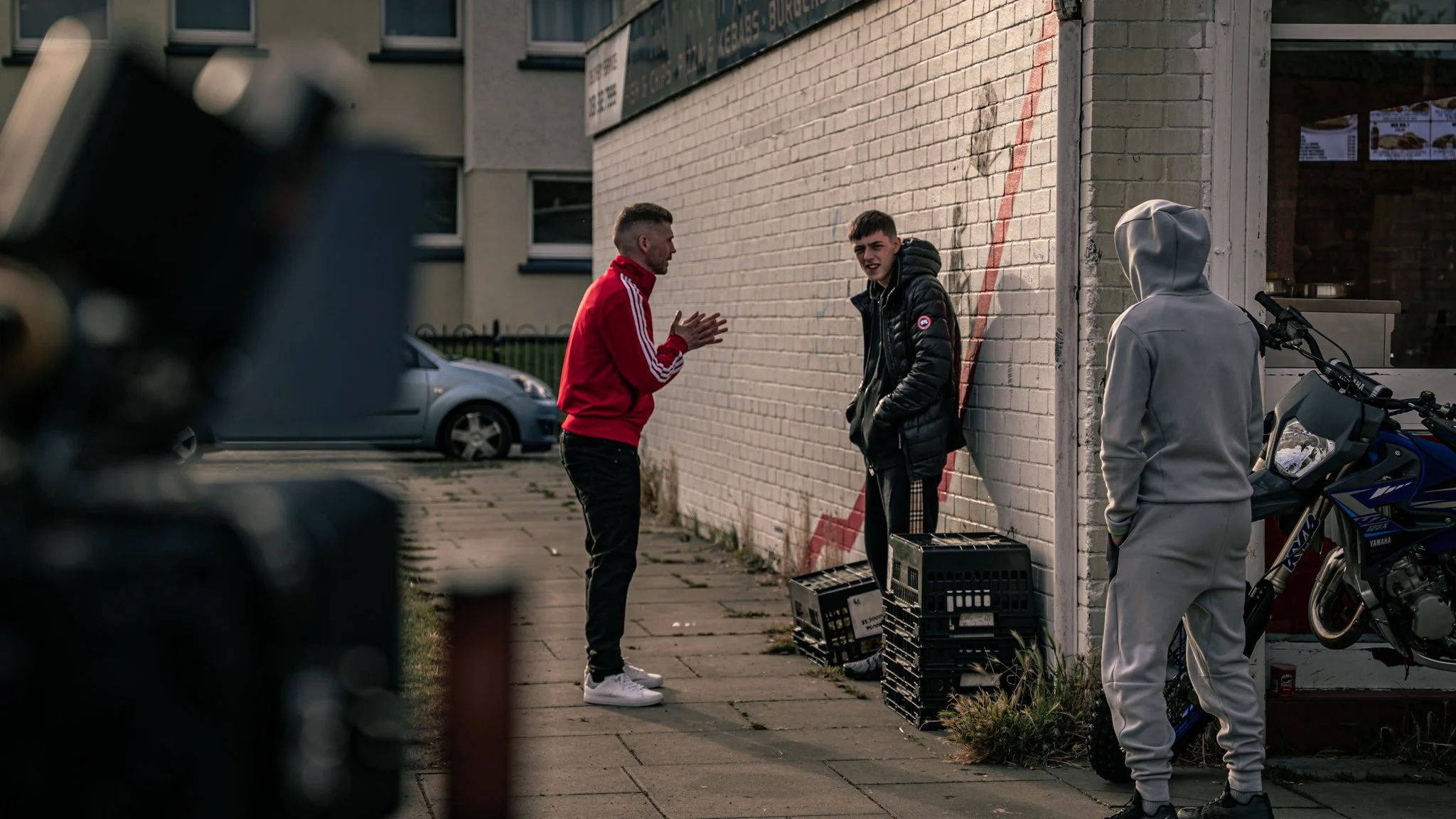 Three young men are standing outside on a sidewalk next to a brick building. One man wearing a red jacket appears to be talking and gesturing to the other two. One of the other men is wearing a black puffer jacket and the third is wearing a gray hoodie with the hood up. There are crates and a motorcycle nearby, and a car parked in the background.