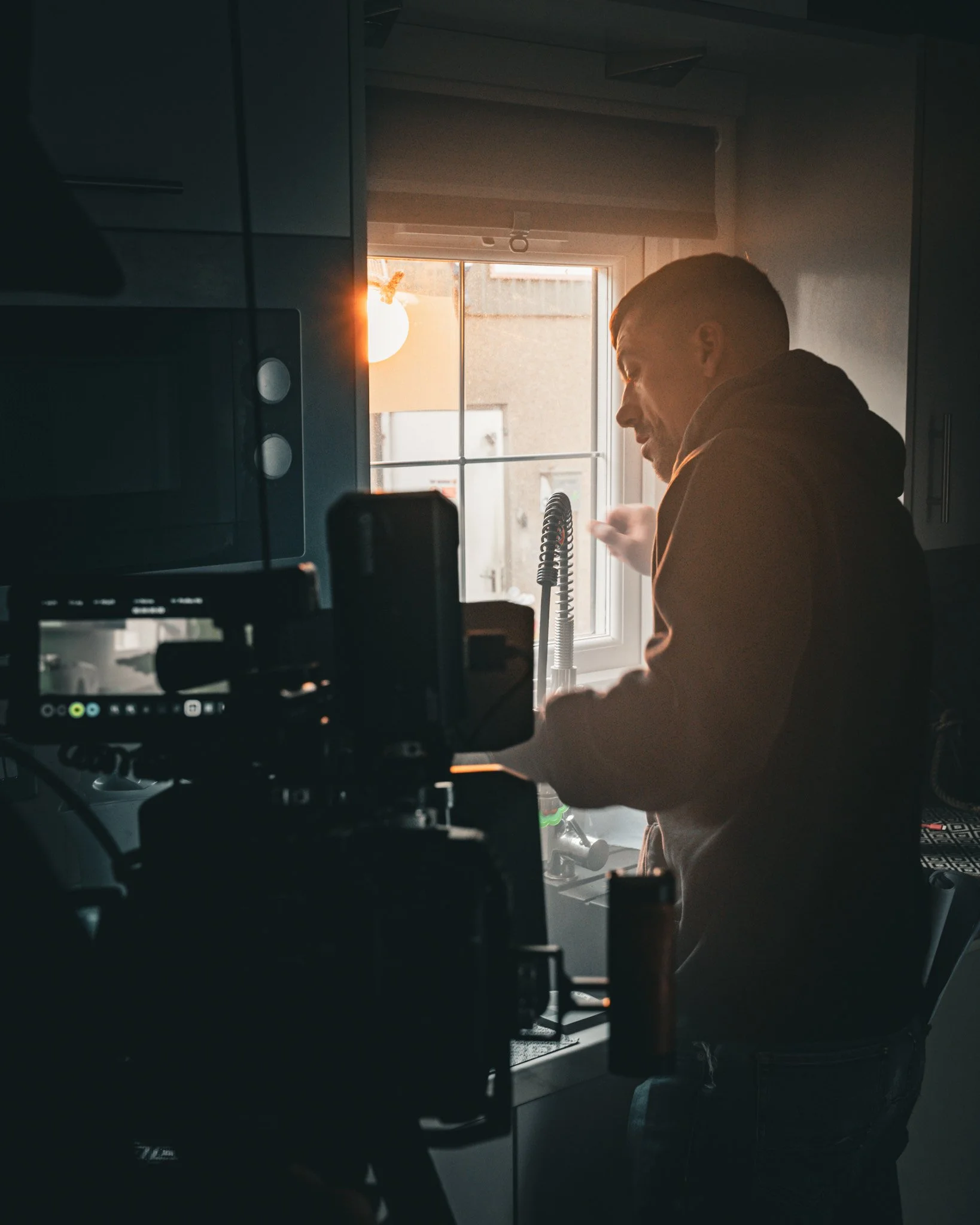 A person in a kitchen standing by a window, with sunlight shining in, wearing a hoodie and looking down at something in their hands. Kitchen appliances and a camera equipment are visible in the foreground.