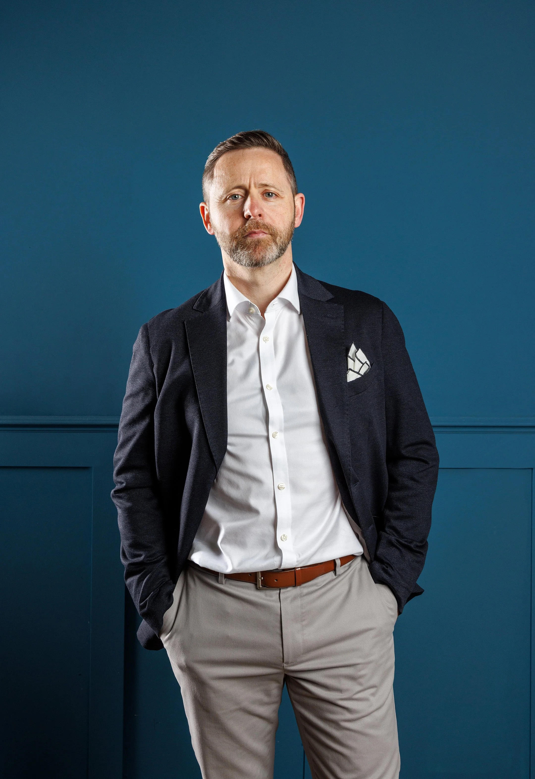 A man in a white shirt, dark blazer, and beige pants stands against a blue wall with paneling. He has short brown hair, a beard, and has a serious expression.