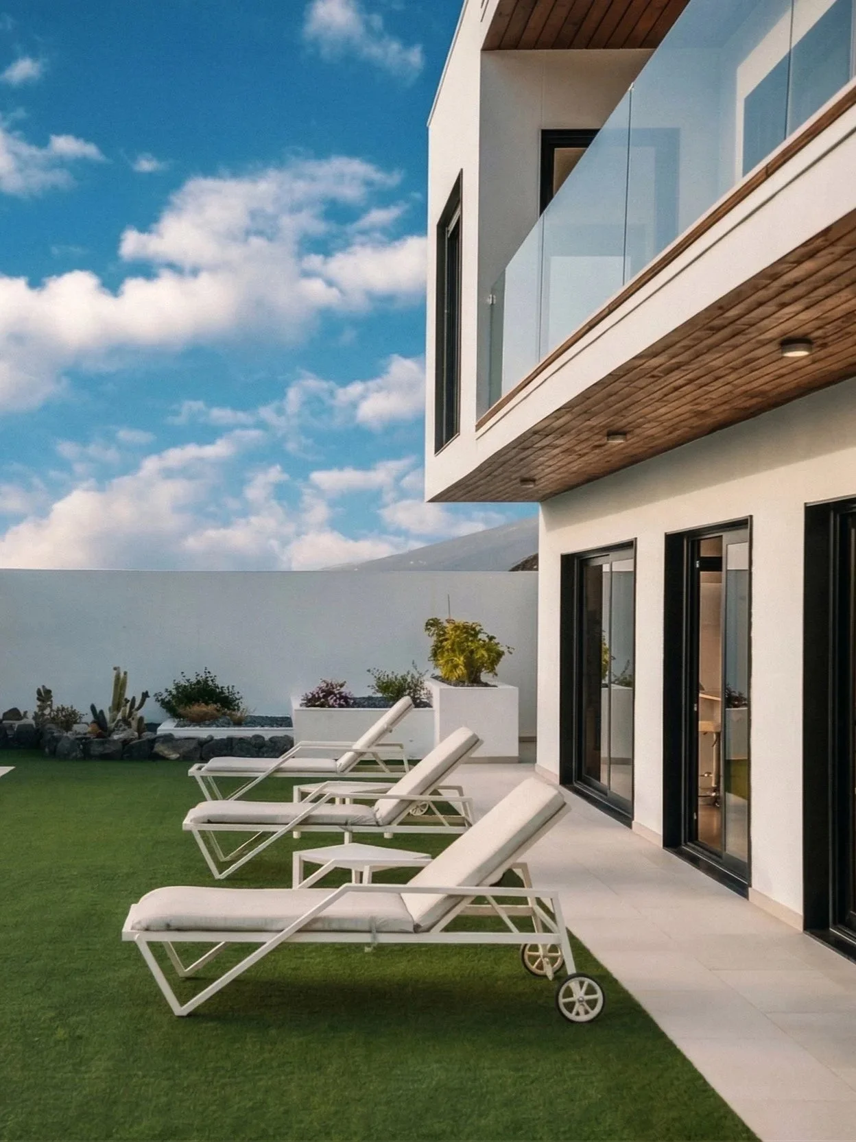 Vista de una terraza con sillones reclinables blancos junto a una casa moderna blanca con ventanas de piso a techo y un balcón de vidrio, cielo despejado y algunos arbustos decorativos en macetas.