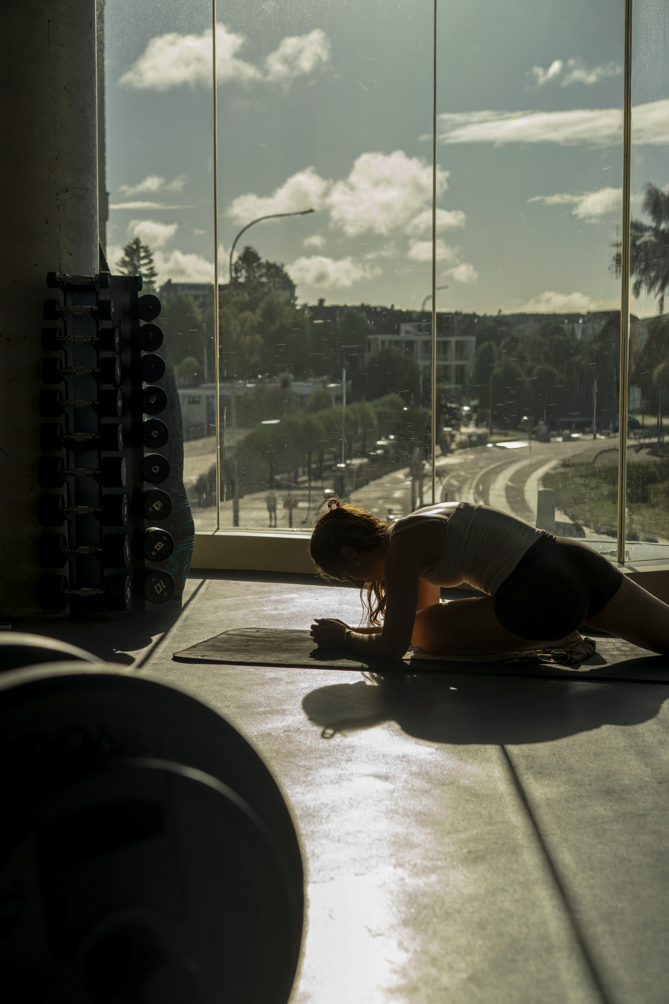 A woman is doing yoga or stretching on a mat in a gym with large windows, sunlight streaming in, overlooking Bondi Beach.
