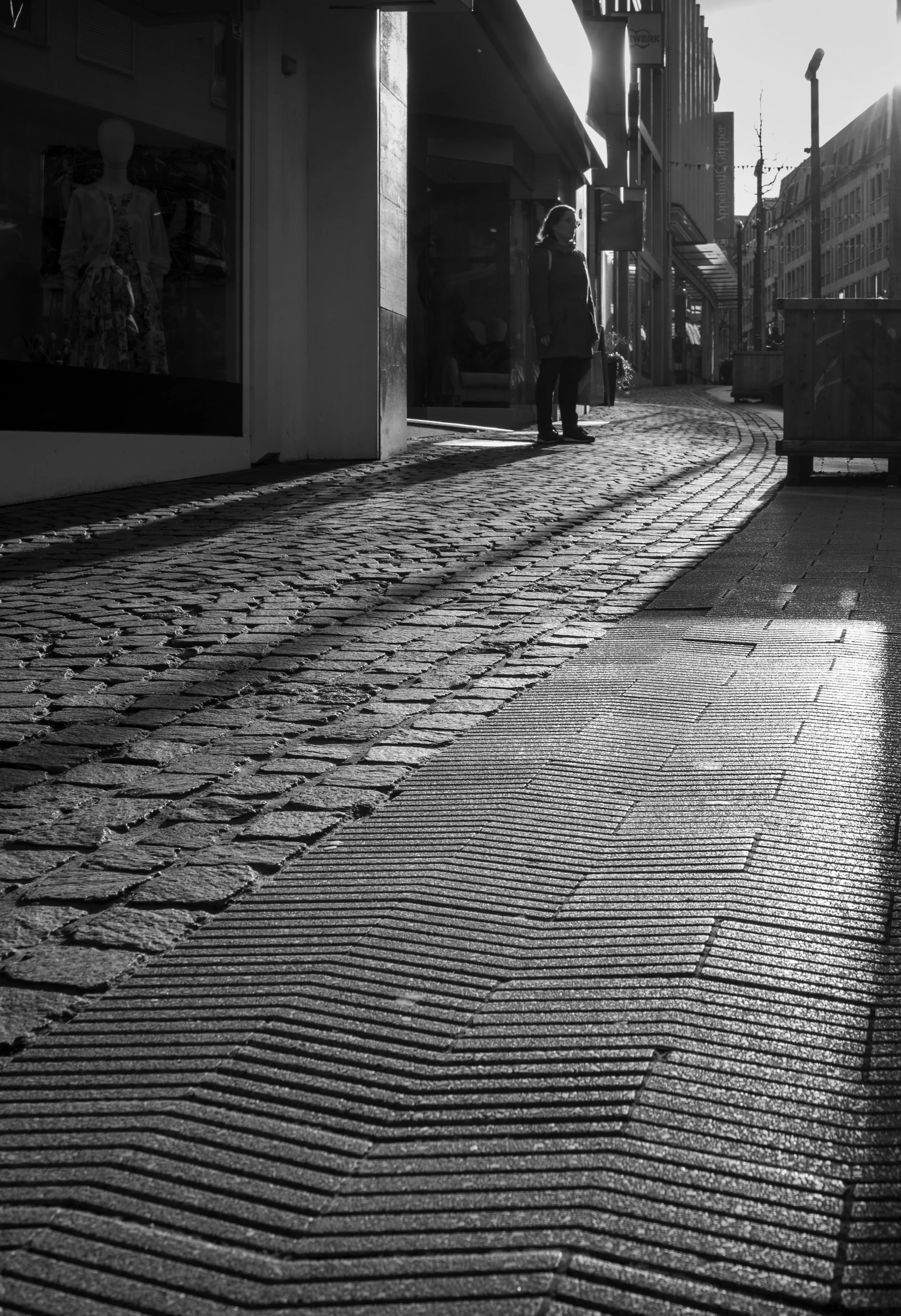Long shadow of a backlit person in the streets of Aachen. In the shopping window a dressmodel watching the scene