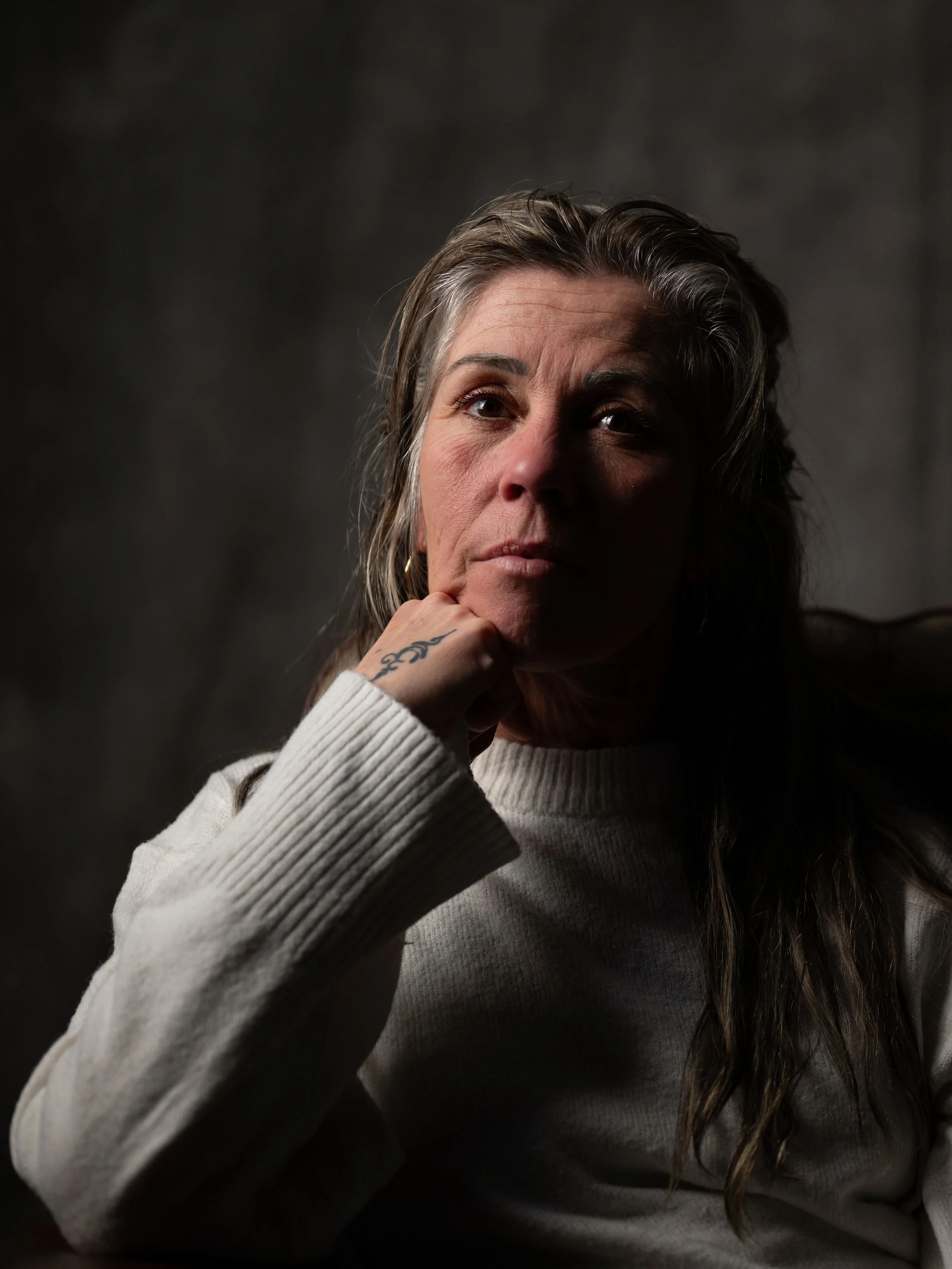 A woman with long brown hair, wearing a white sweater, resting her chin on her hand, in front of a dark background.