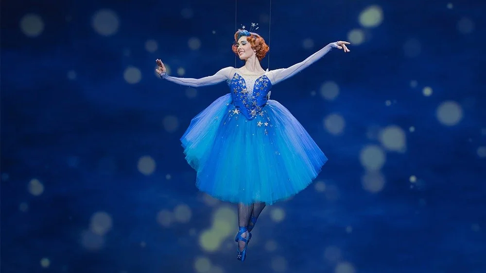 A ballerina in a blue tutu and long-sleeved leotard performing a leap with arms outstretched against a dark blue background with glowing bokeh lights.