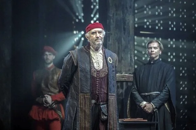 Three people dressed in historical costumes standing on a stage with dramatic lighting and a starry background.
