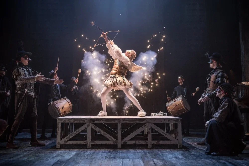 A circus performer dressed as a gladiator standing on a wooden platform, holding a bow and arrow, surrounded by sparks and smoke, with four performers in black costumes and masks playing drums and percussion in a dark theater.