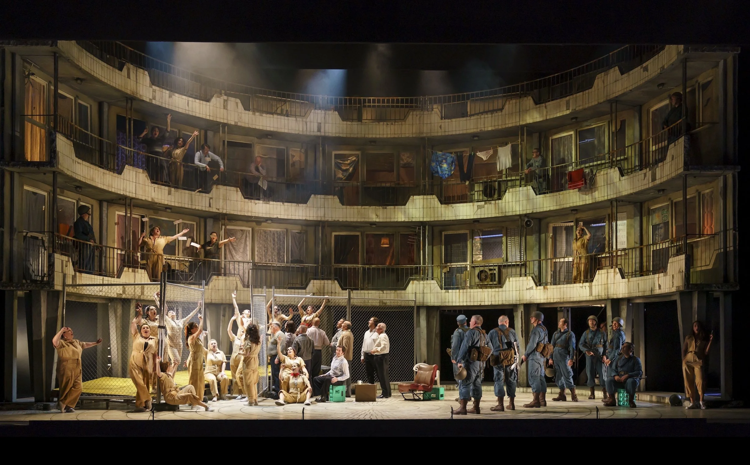 Scene from a theatrical performance with multiple actors on stage and on balconies, some wearing costumes and military uniforms, with a crowd of spectators watching in front.