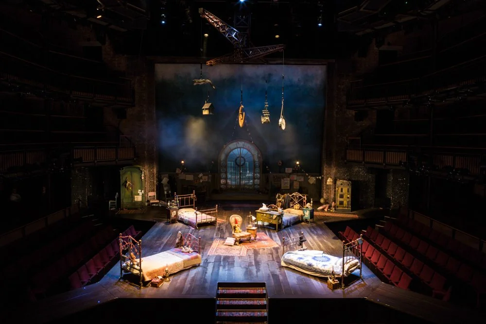 Empty stage set for a theatrical play with vintage furniture, beds, and hanging props, illuminated with stage lights in a theater with balconies.