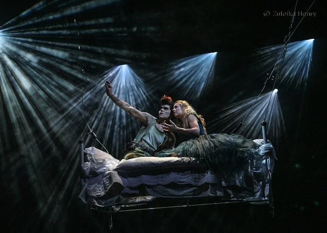 Two performers on a bed suspended in the air during a theatrical scene, illuminated by dramatic spotlighting and stage lights.