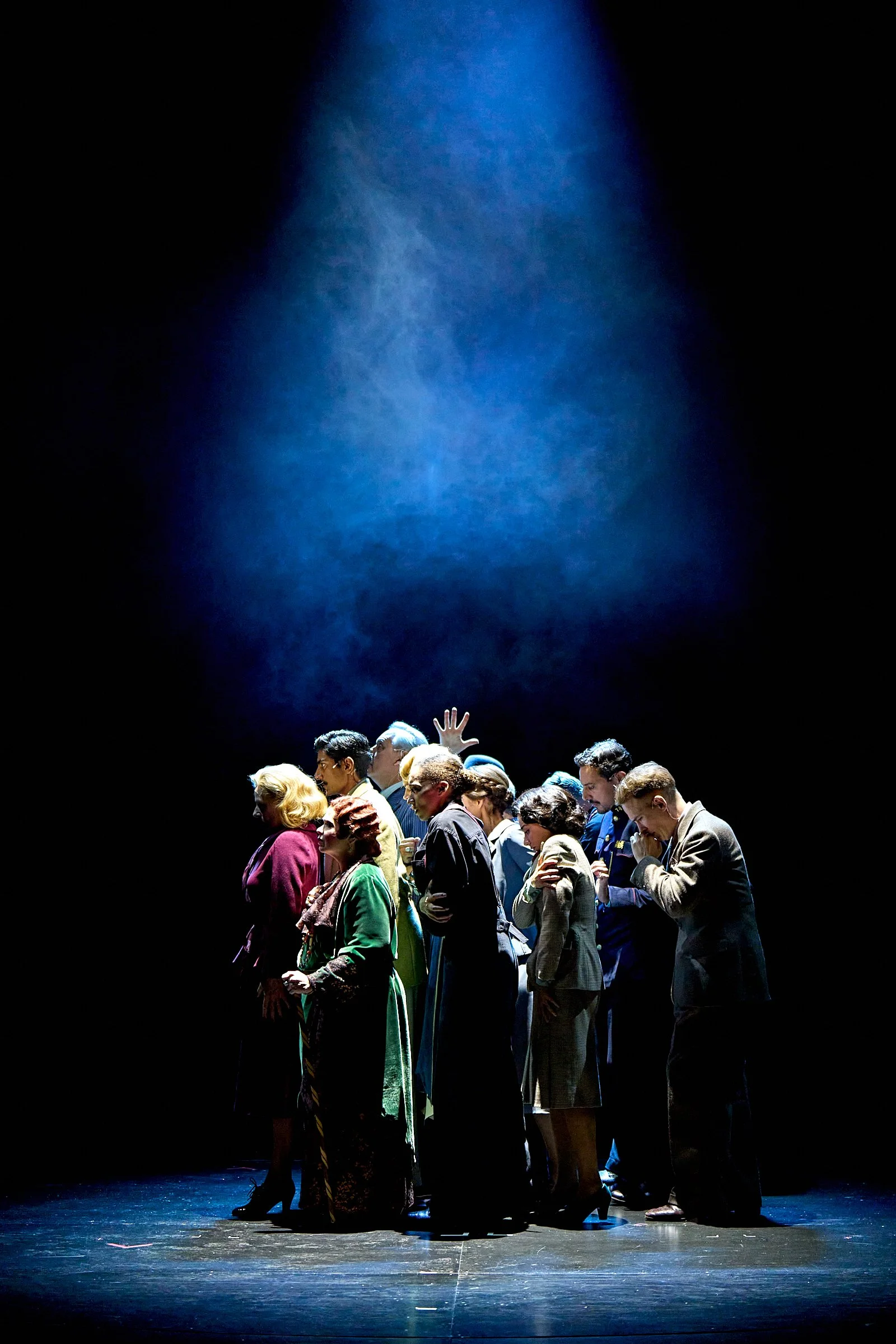 A group of diverse people standing closely together on a dark stage, illuminated by a spotlight from above, with some heads bowed and hands clasped in prayer or reflection.