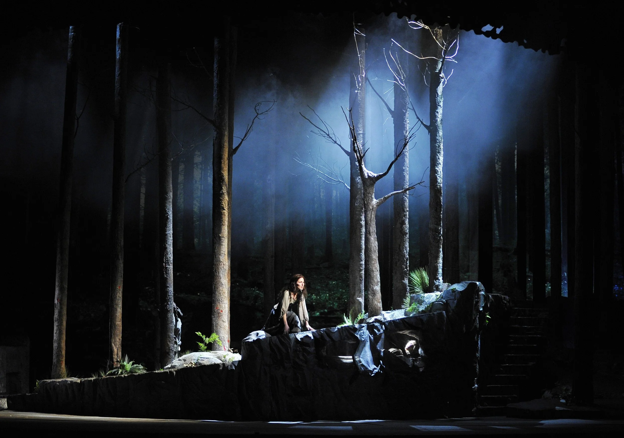 A woman sitting on rocks on a stage with a forest backdrop, illuminated by blue and white lights, creating a mystical atmosphere.