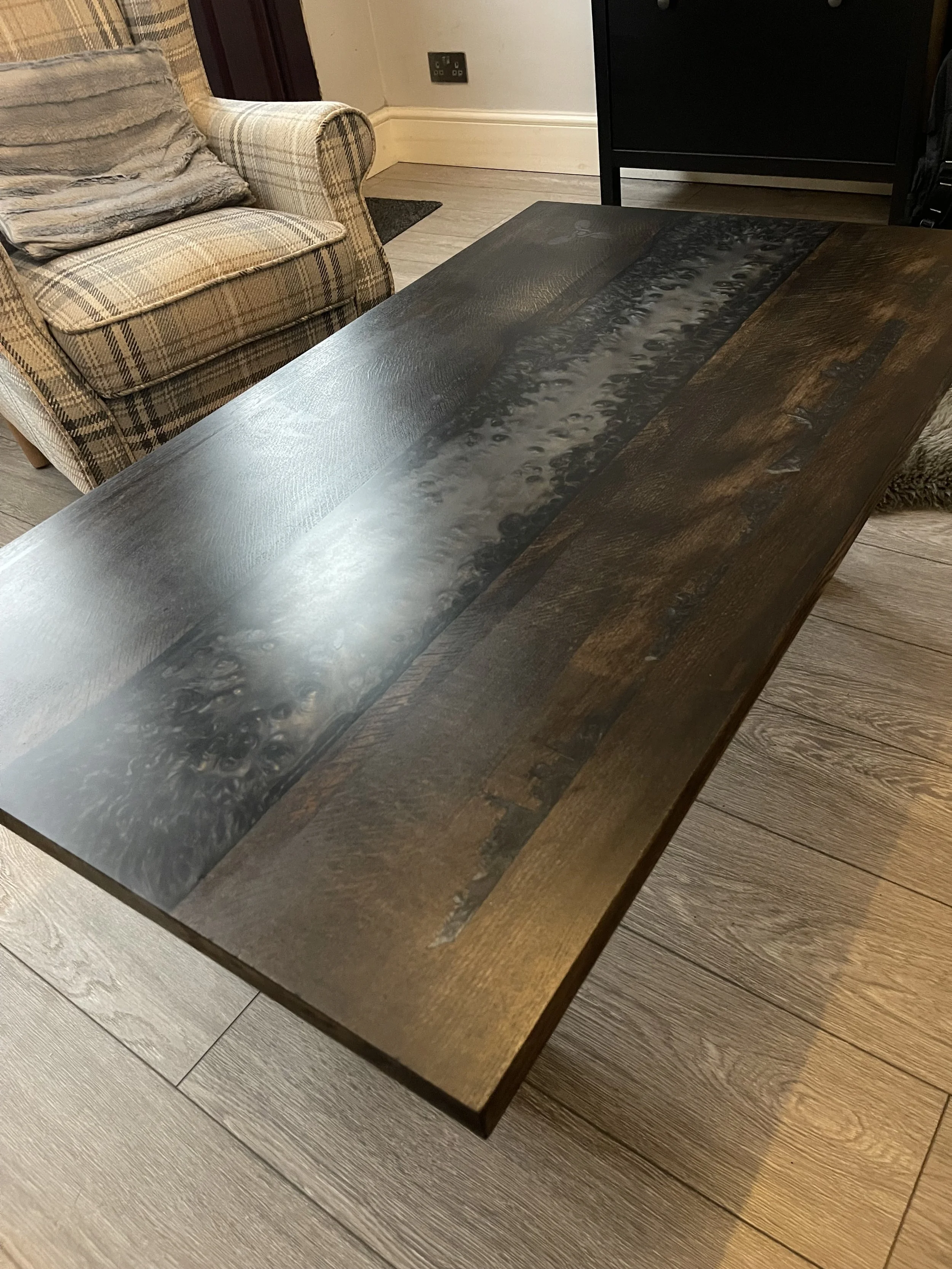A wooden table with a dark finish and a decorative resin inlay running through the center, positioned in a living room near an armchair and a cabinet.