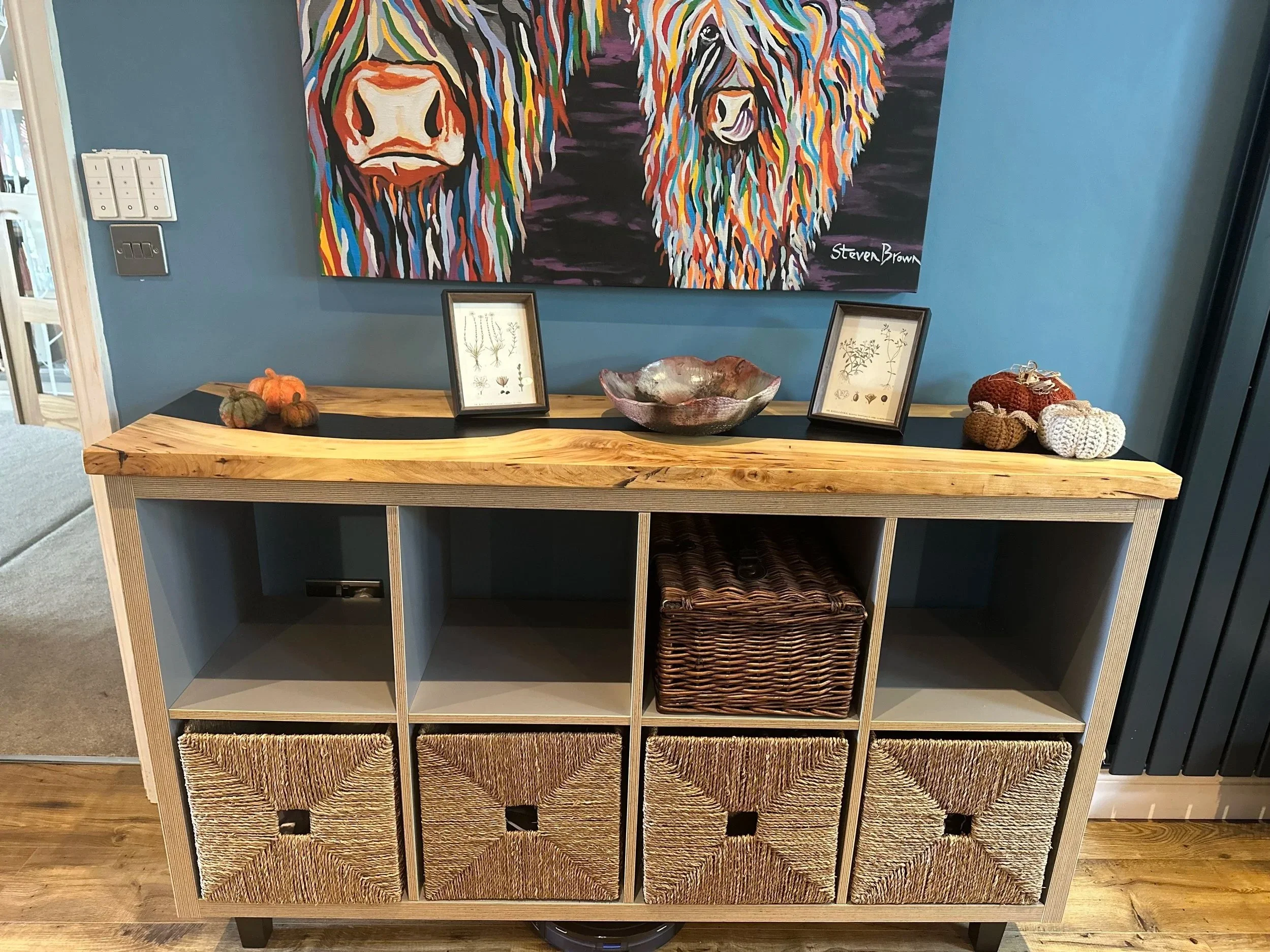 A wooden sideboard with woven baskets on the bottom, a decorative bowl, framed pictures, and small pumpkins on top, against a blue wall with a colorful painting of two bison.